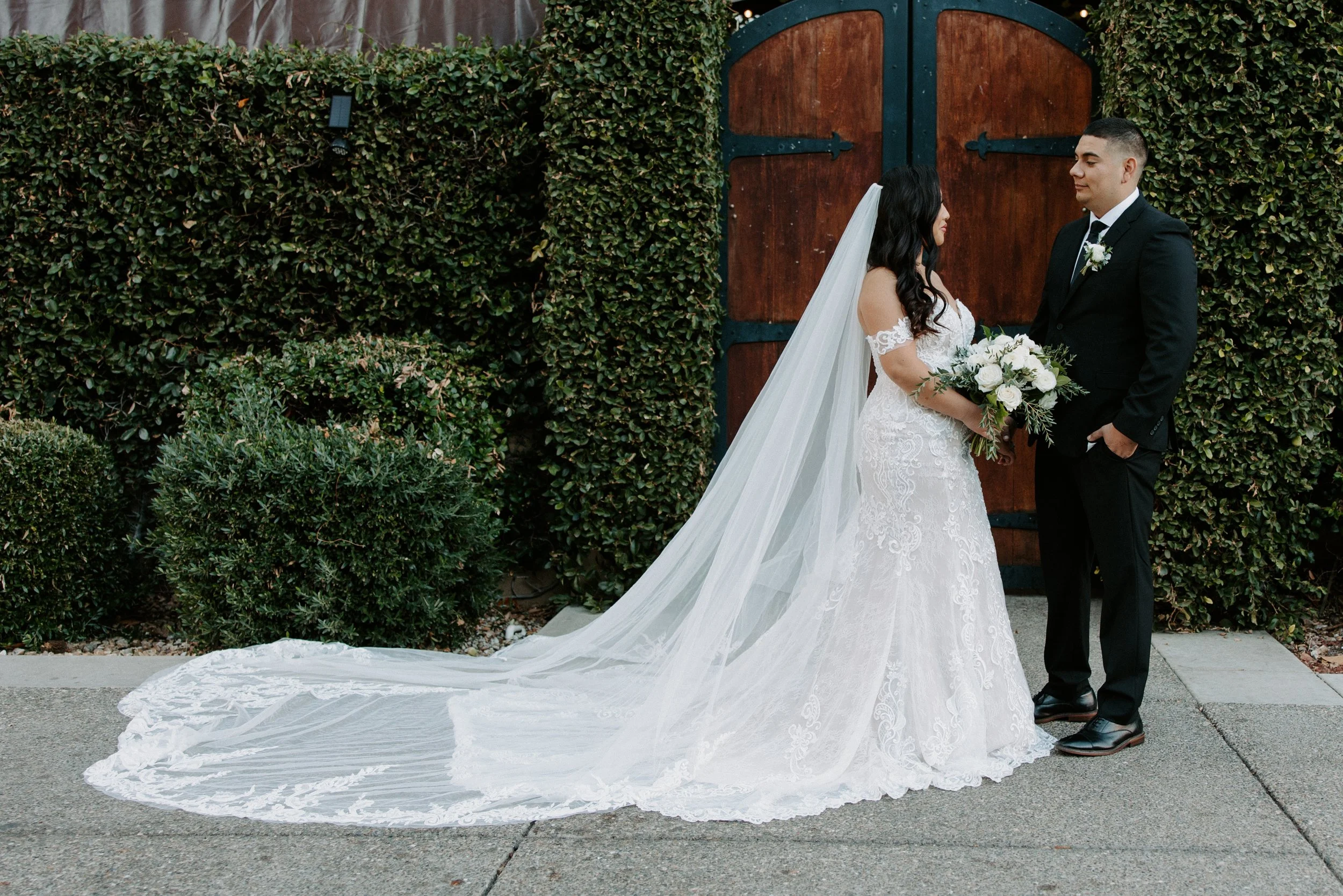 Bride and groom standing face to face, holding hands during their wedding outside, with lush green hedge and wooden gate behind them.
