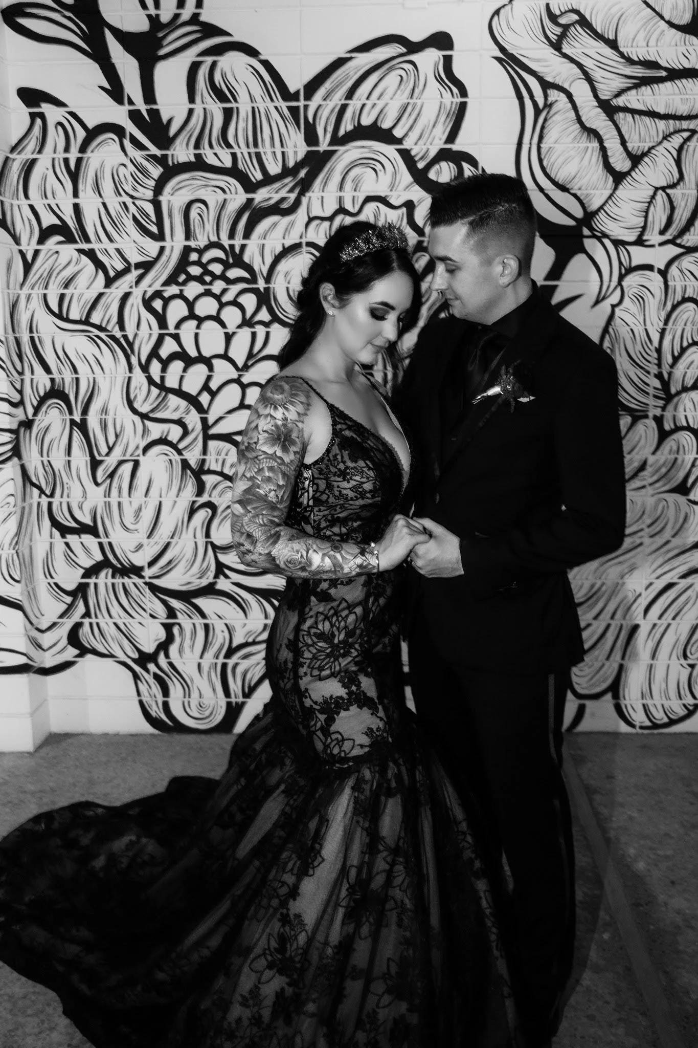 A black and white photo of a couple dressed in formal attire, standing close and holding hands in front of a wall with an artistic floral mural.