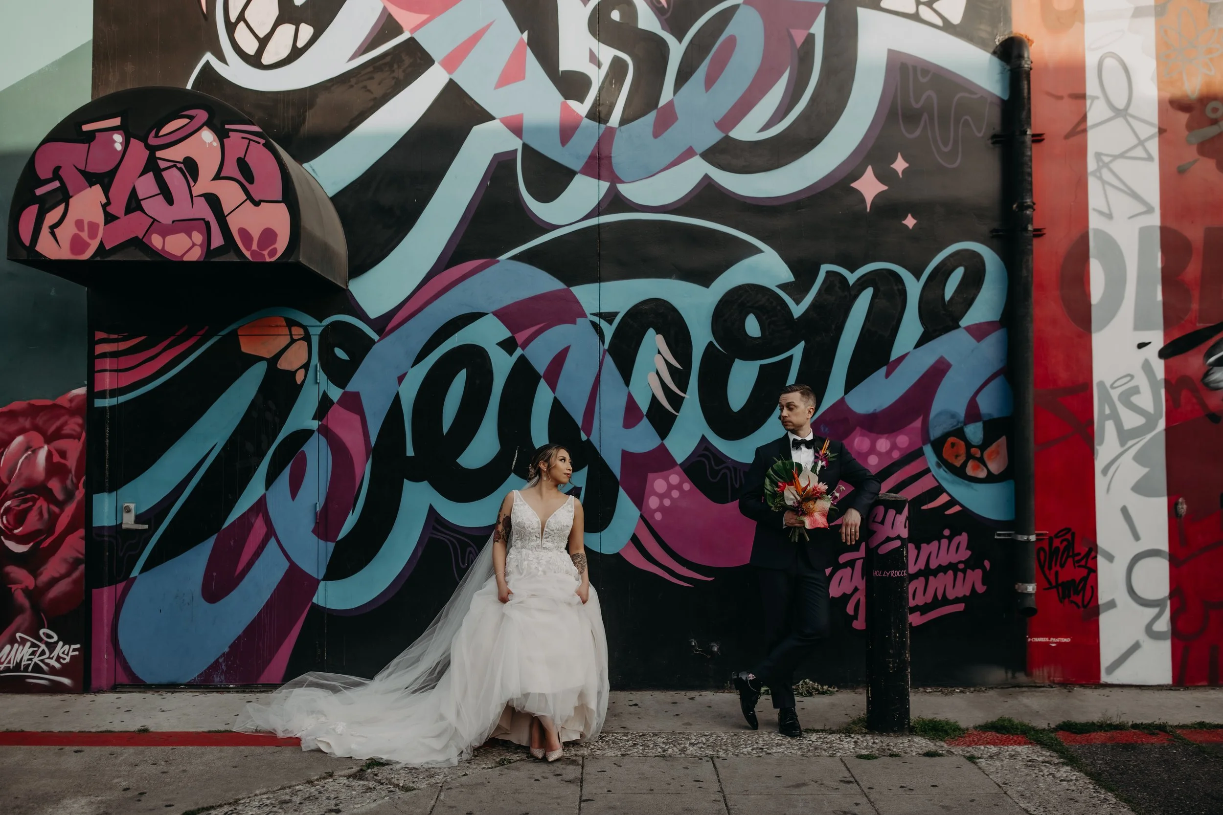 A bride in a white wedding dress and a groom in a black tuxedo with a bouquet, standing in front of a colorful graffiti wall.