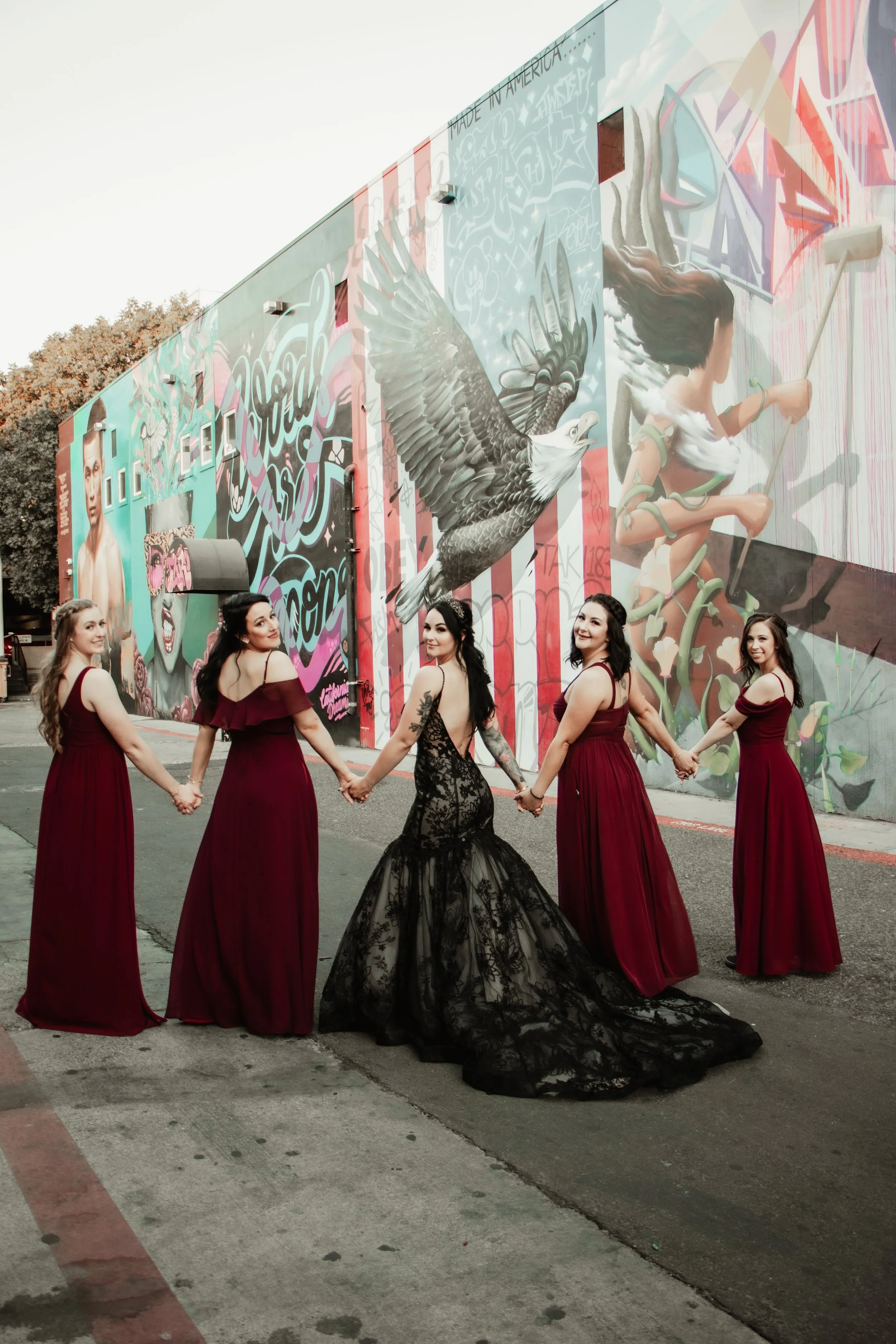 Five women in matching long burgundy dresses holding hands and standing in front of a colorful graffiti mural on the wall.