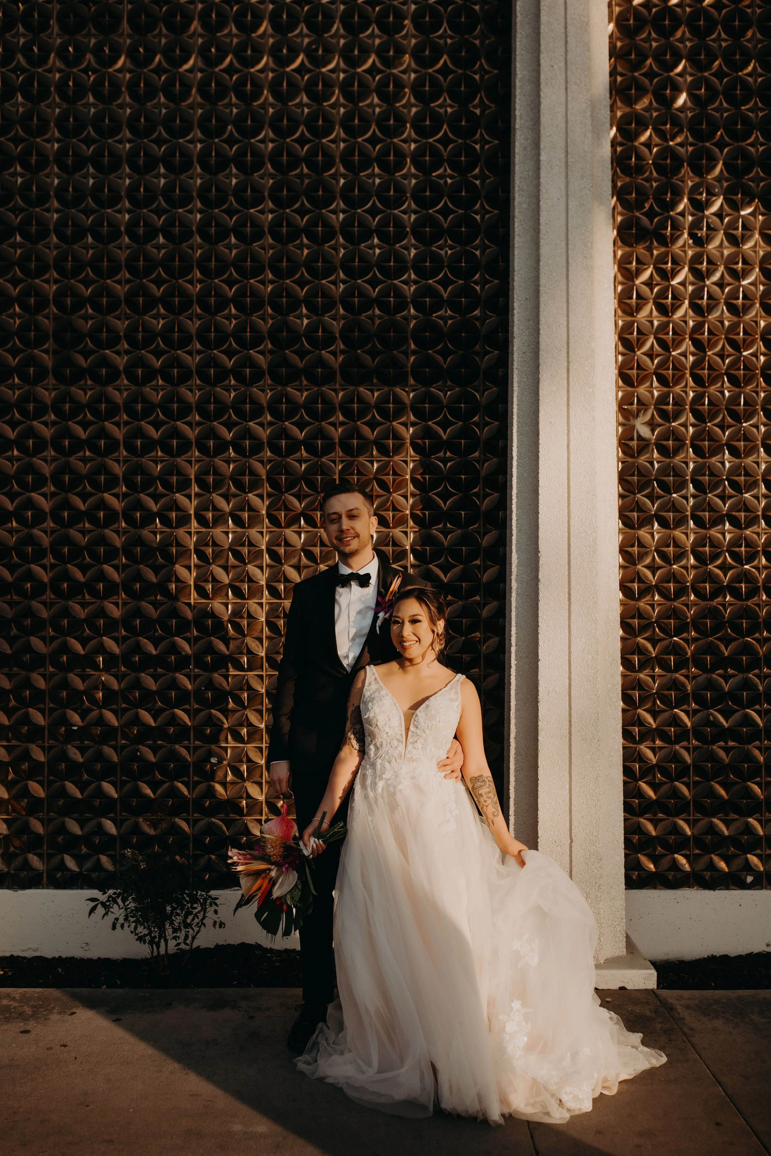A bride and groom standing outdoors in front of a decorative wall, with the bride holding a bouquet and wearing a white wedding gown, and the groom in a black tuxedo.