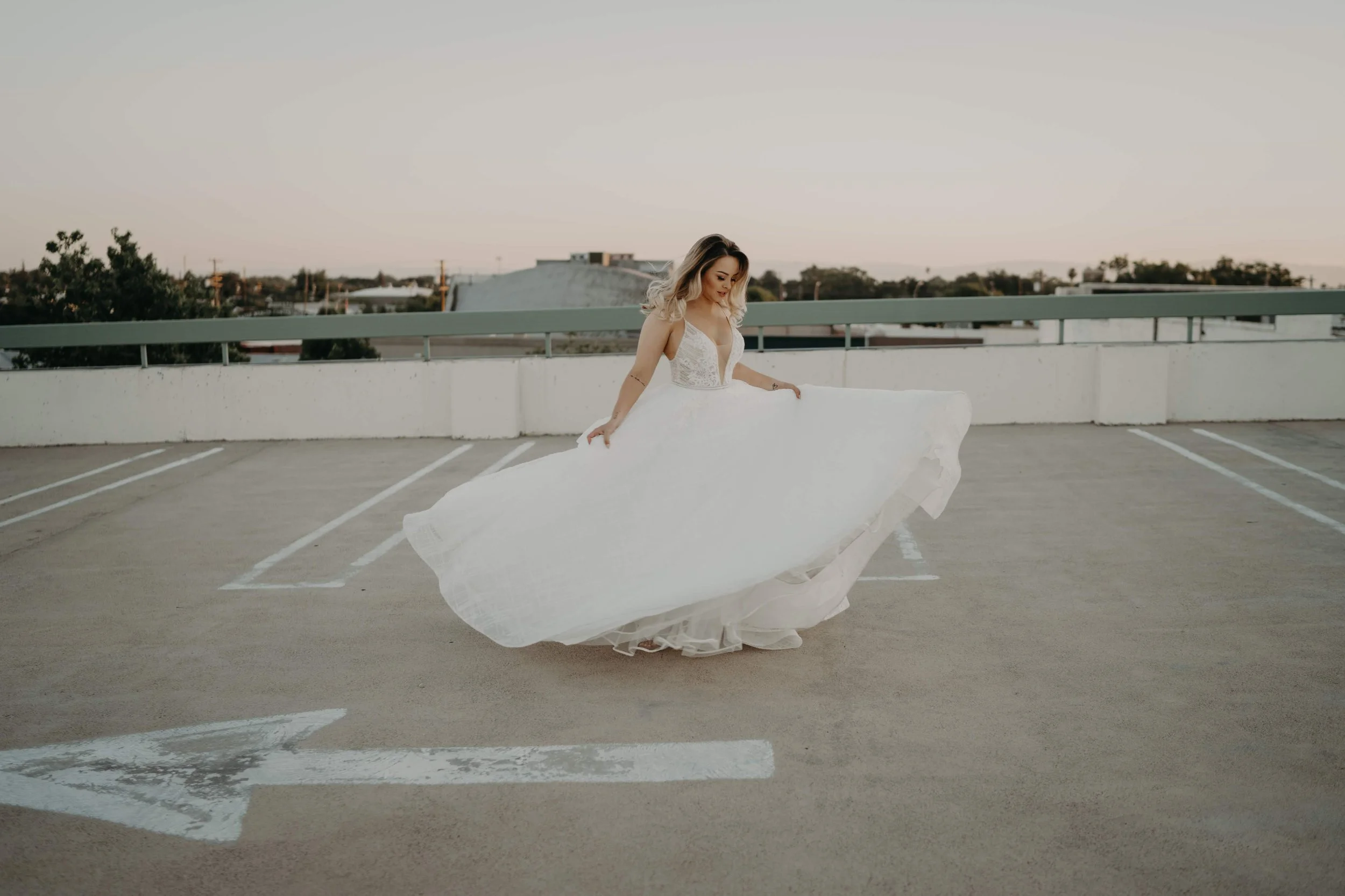 A woman in a wedding dress standing on a rooftop parking lot, holding her dress out to show its fullness, with a cityscape in the background at sunset.