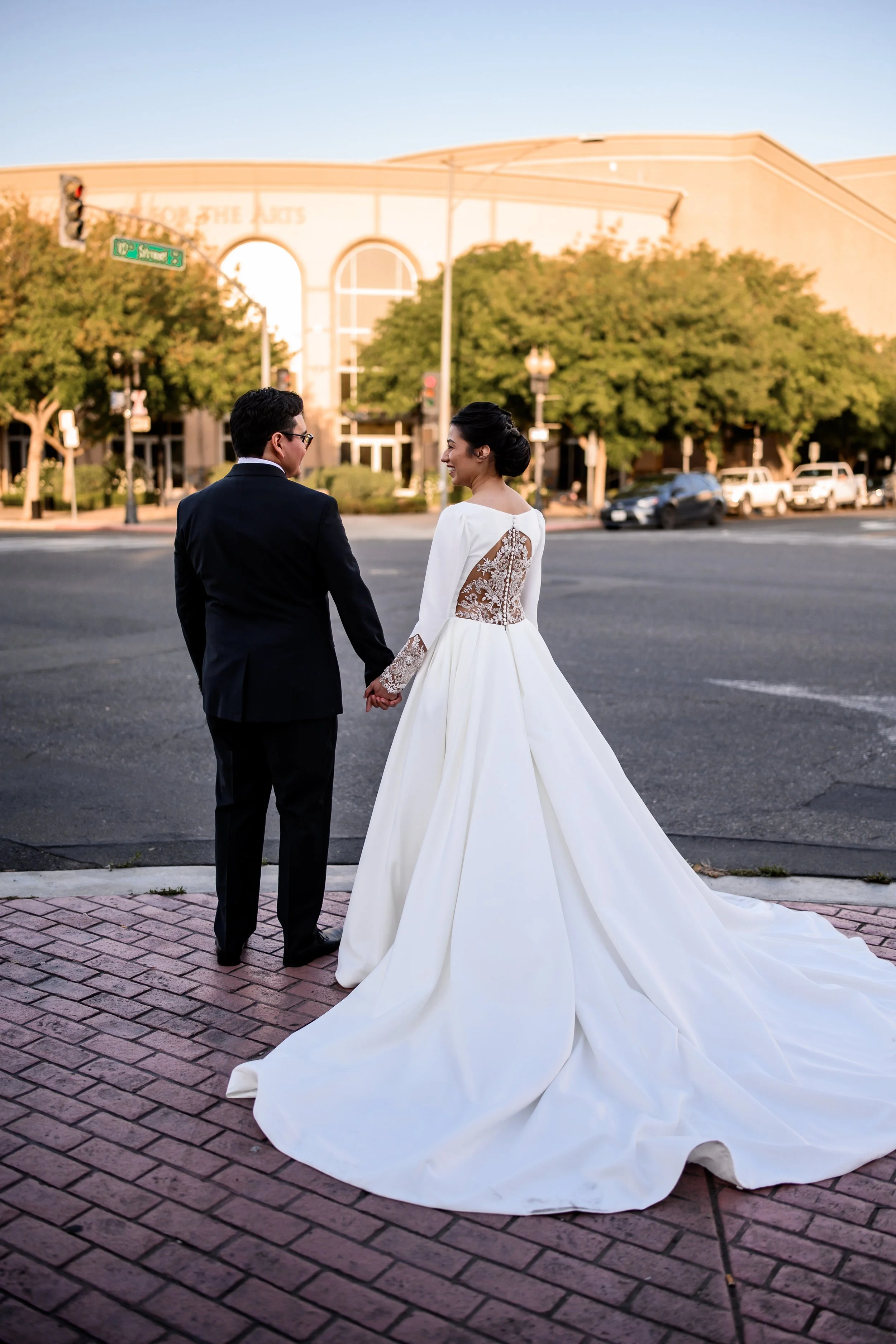 A bride and groom holding hands on a city sidewalk, dressed in wedding attire, with the bride wearing a long white gown with lace back details and the groom in a black tuxedo, during sunset.