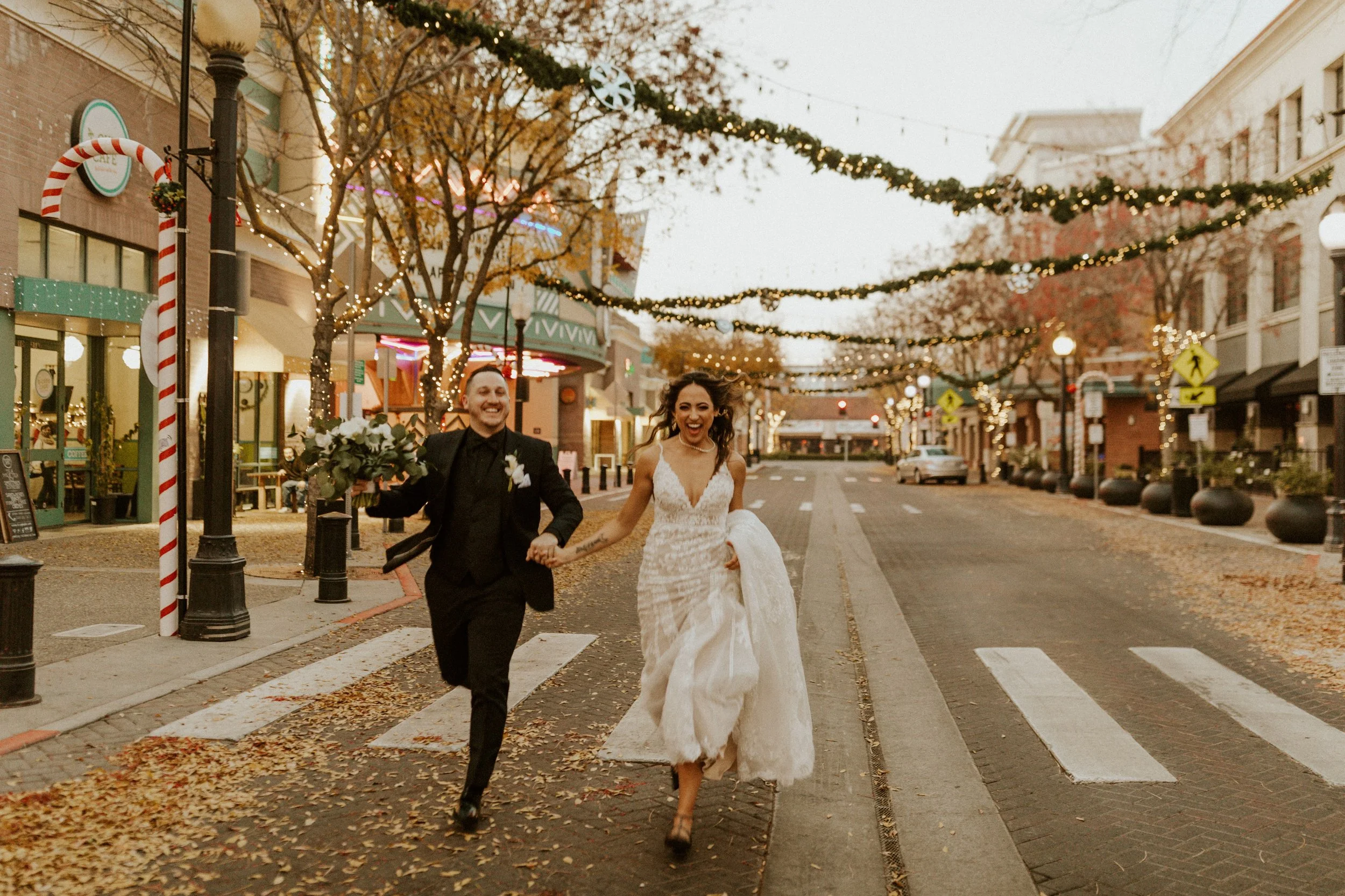 A newlywed couple running hand in hand down a decorated city street with festive string lights and autumn leaves on the ground.