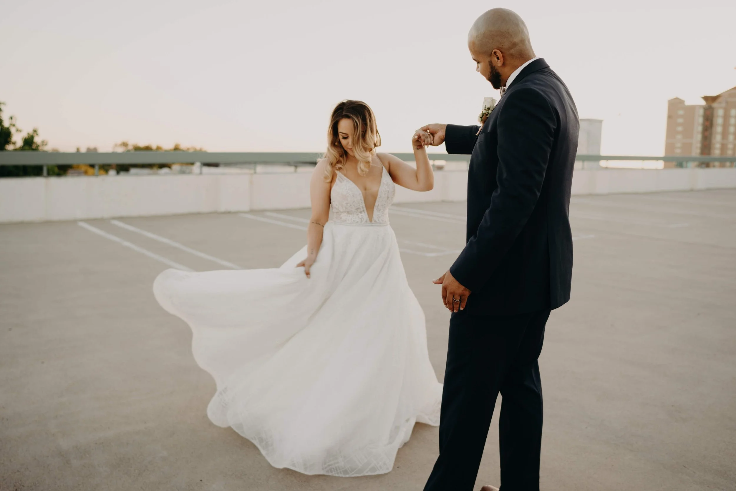 A bride in a white wedding dress dancing with a groom in a black suit on a parking garage rooftop during sunset.