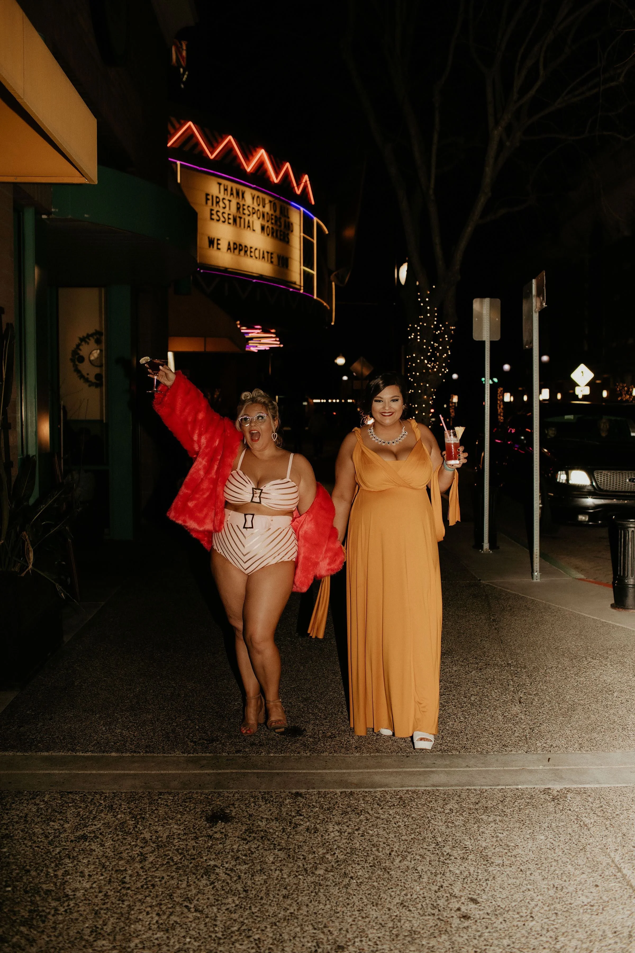 Two women walking on a city sidewalk at night, dressed in glamorous outfits, both holding drinks and smiling.