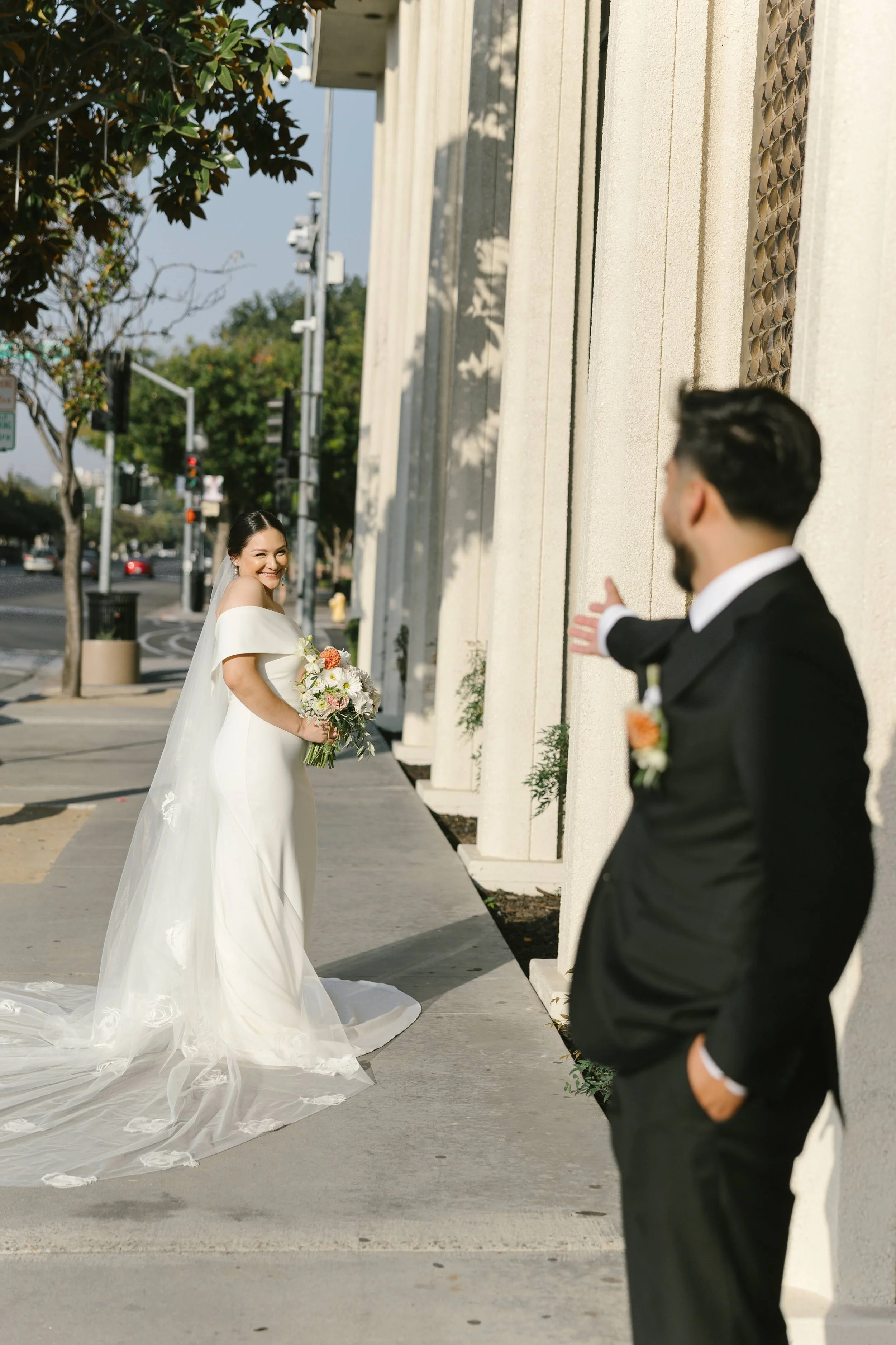 A bride in a white wedding dress holding a bouquet, smiling and looking at her groom, who is in a black tuxedo, on a city sidewalk near a building with columns.