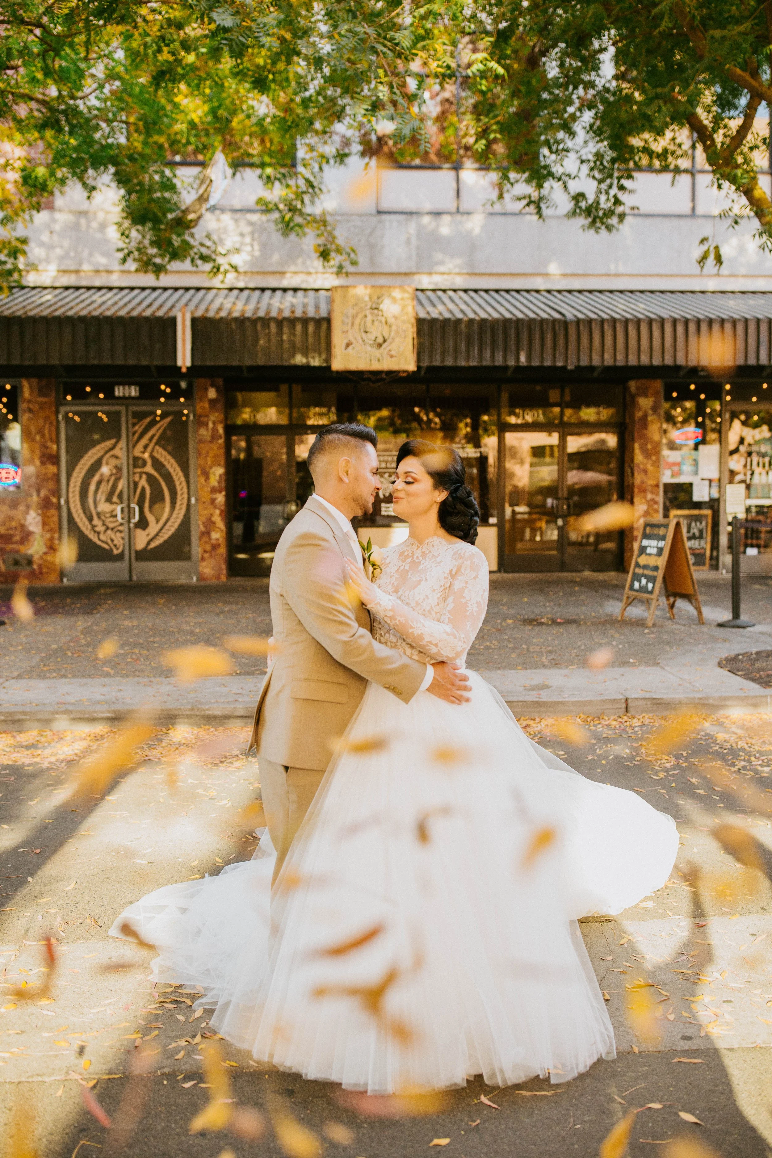 A bride and groom dancing outside a restaurant, holding each other closely, with leaves falling around them and autumn trees in the background.
