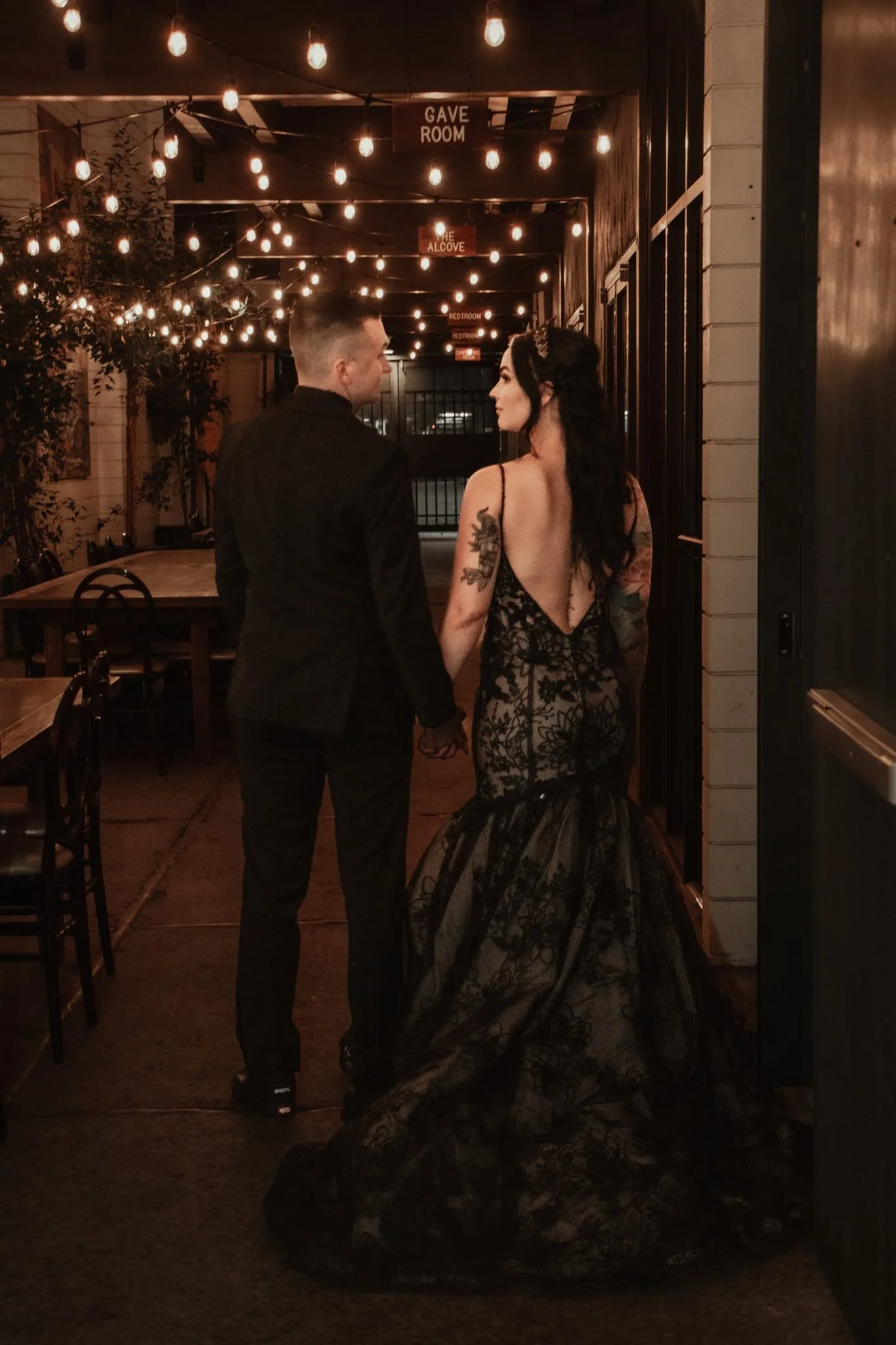 A man in a black suit and a woman in a black lace dress walking hand in hand through a dimly lit outdoor area decorated with string lights at night.