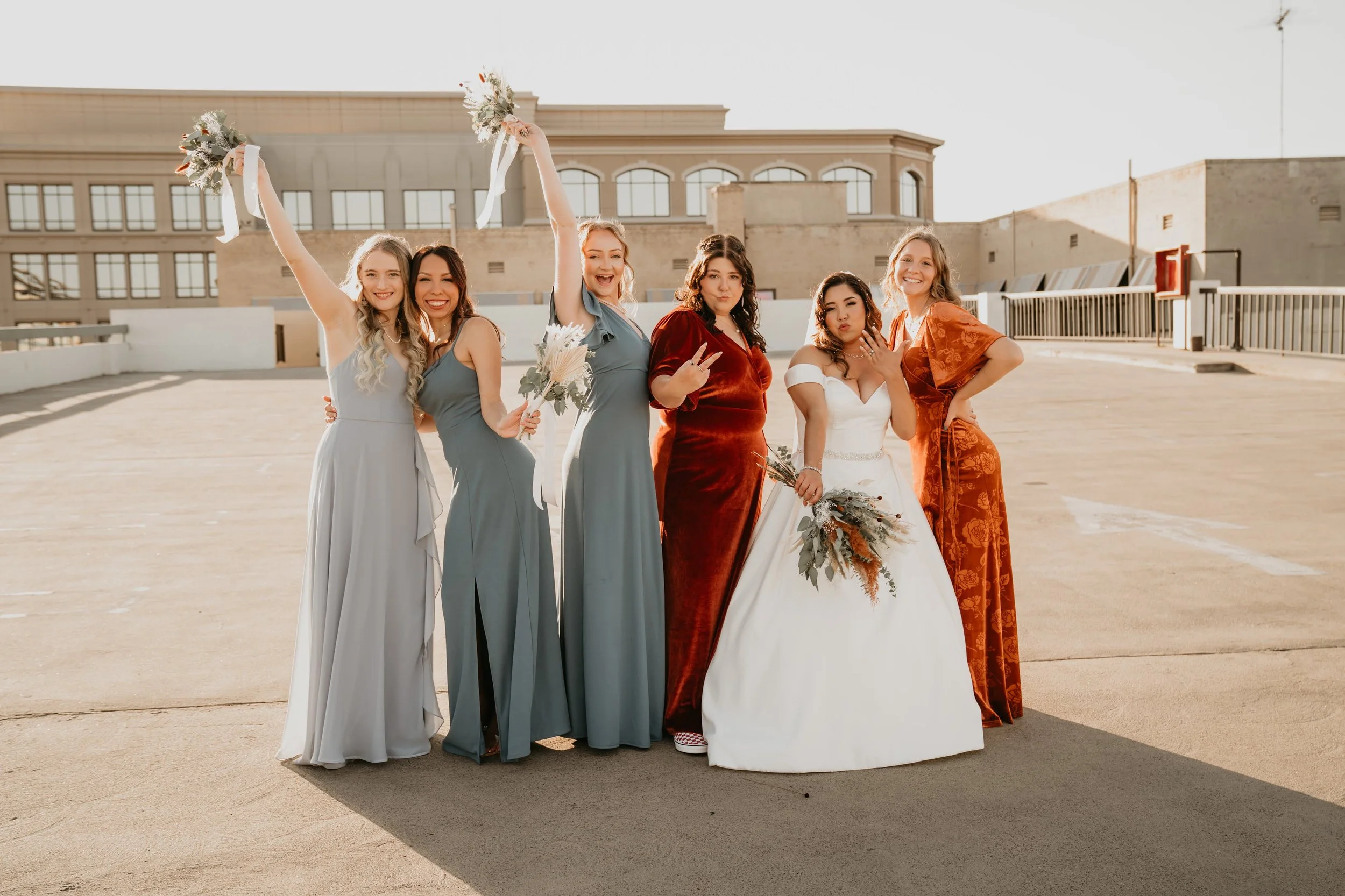 Six women dressed in bridesmaid and wedding gowns, posing and smiling on a rooftop parking deck with a city building in the background during late afternoon.