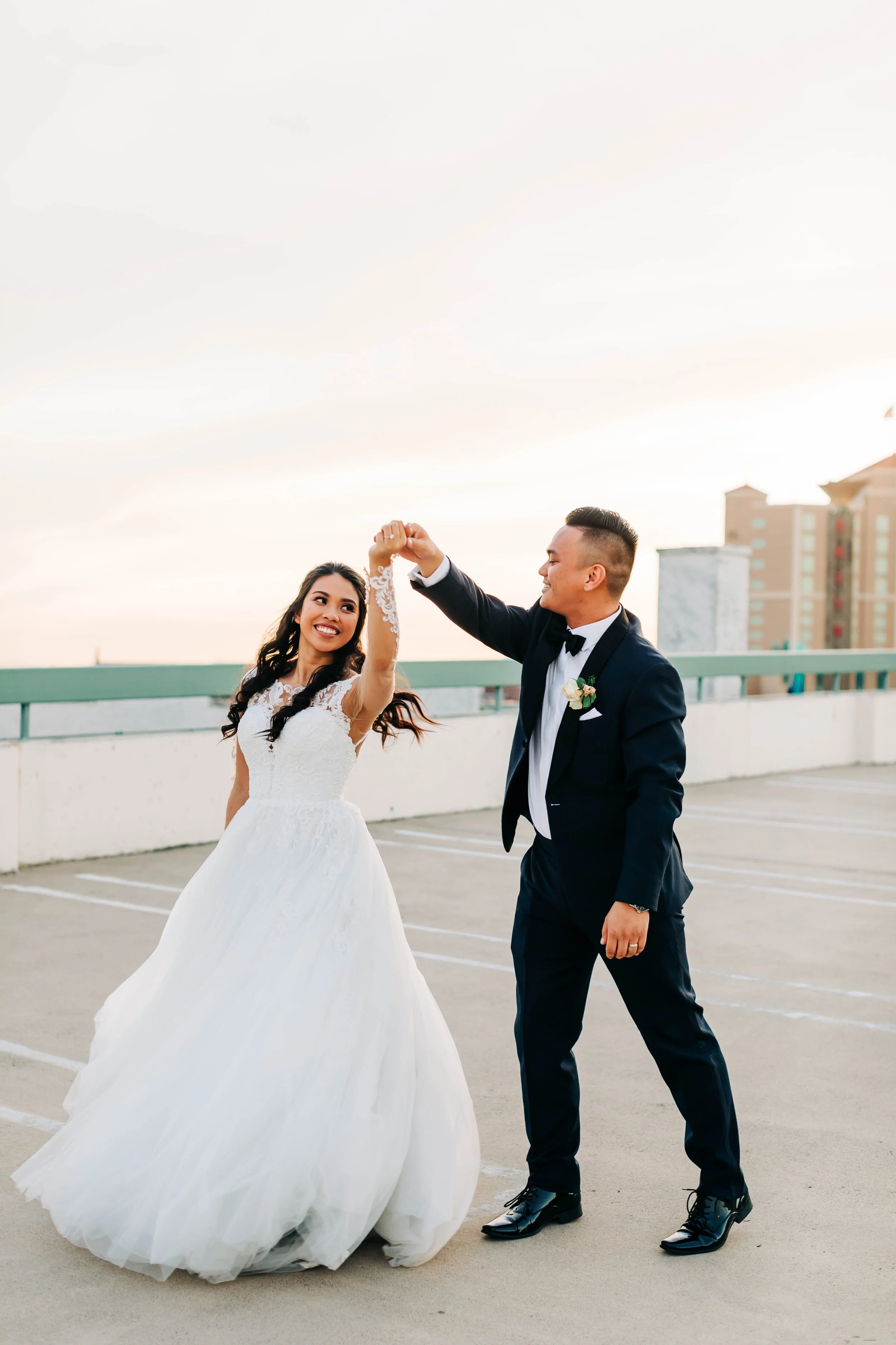 A bride and groom dancing on a rooftop parking lot during sunset, the bride wearing a white wedding gown and the groom in a black tuxedo with a bowtie, smiling and holding hands.