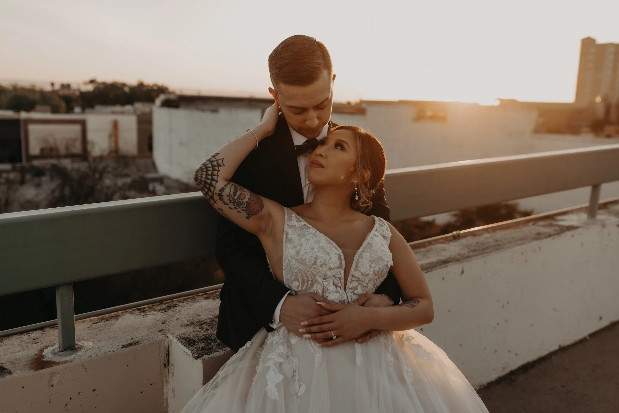 A bride and groom on a rooftop at sunset, embracing and gazing into each other's eyes. The bride is wearing a white lace wedding dress with a sweetheart neckline, and the groom is in a black tuxedo with a bow tie. The bride has tattoos on her arms an
