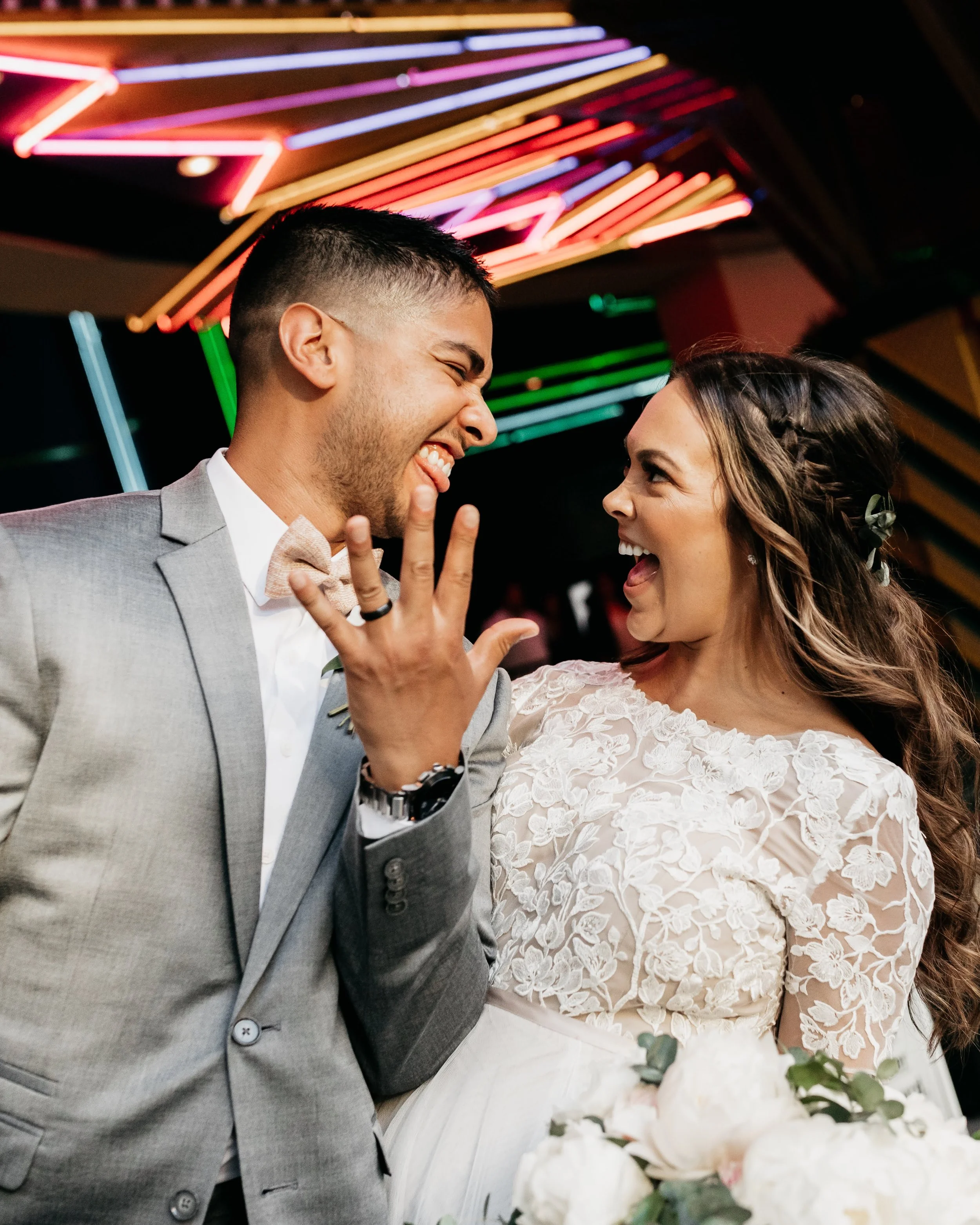A joyful couple at their wedding celebration, smiling and laughing with colorful neon lights in the background.