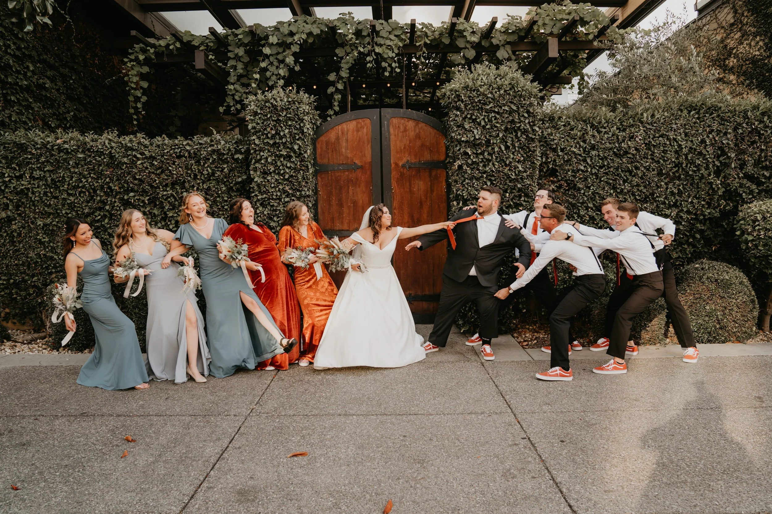 A playful wedding photo featuring a bride and groom in the center, with bridesmaids on one side and groomsmen on the other, all engaged in humorous tug-of-war over the groom's tie, set against a large wooden door and lush greenery backdrop.