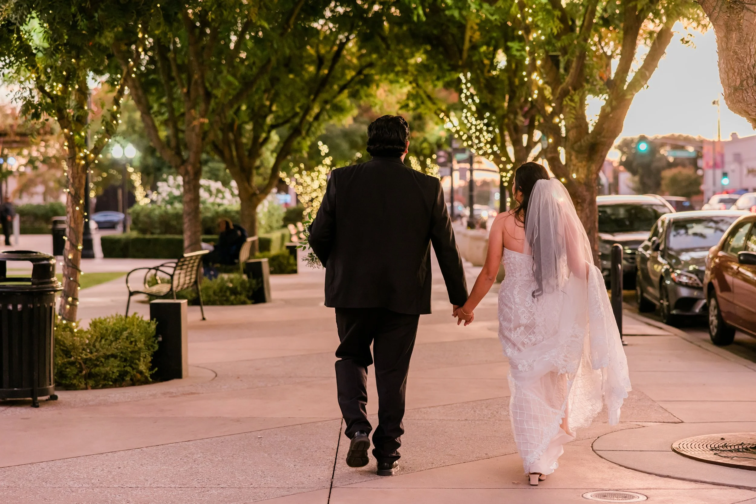 A couple in wedding attire walking hand-in-hand on a decorated city sidewalk at sunset.