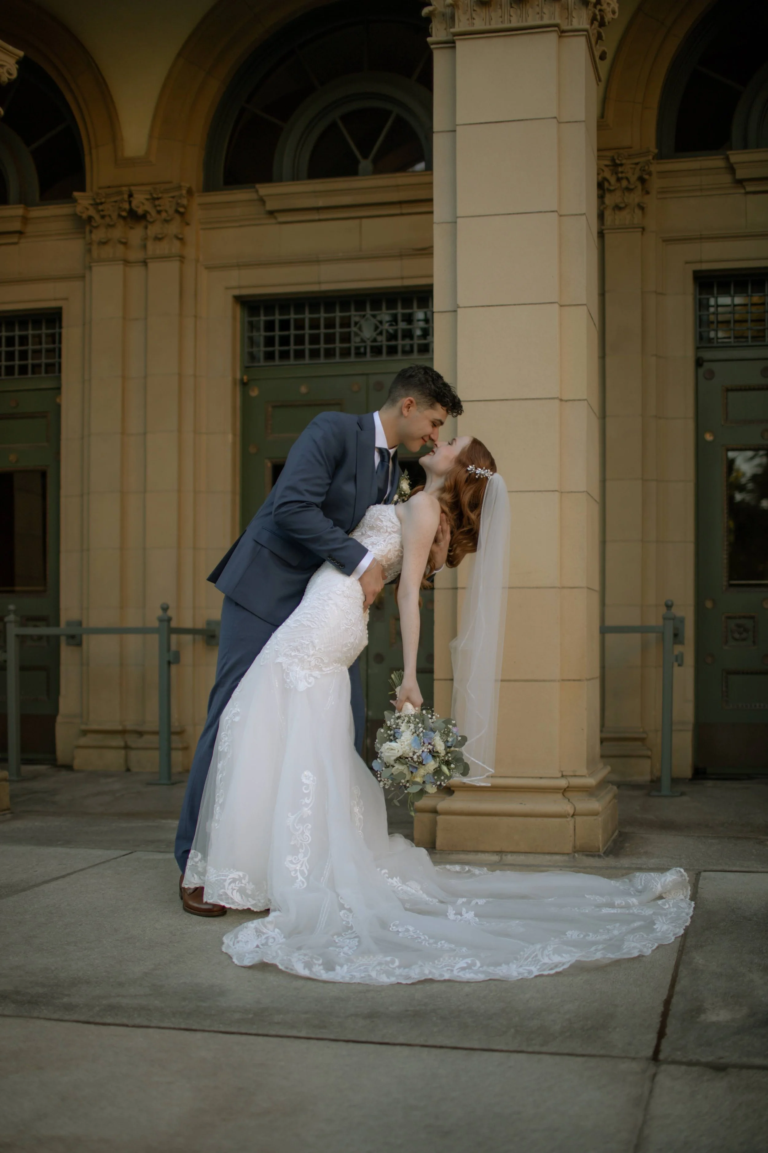 Bride and groom sharing a dance outdoors, with the groom dipping the bride. The bride is holding a bouquet and wearing a white lace wedding gown with a veil, while the groom is in a dark suit. They are in front of a historic building with arched wind