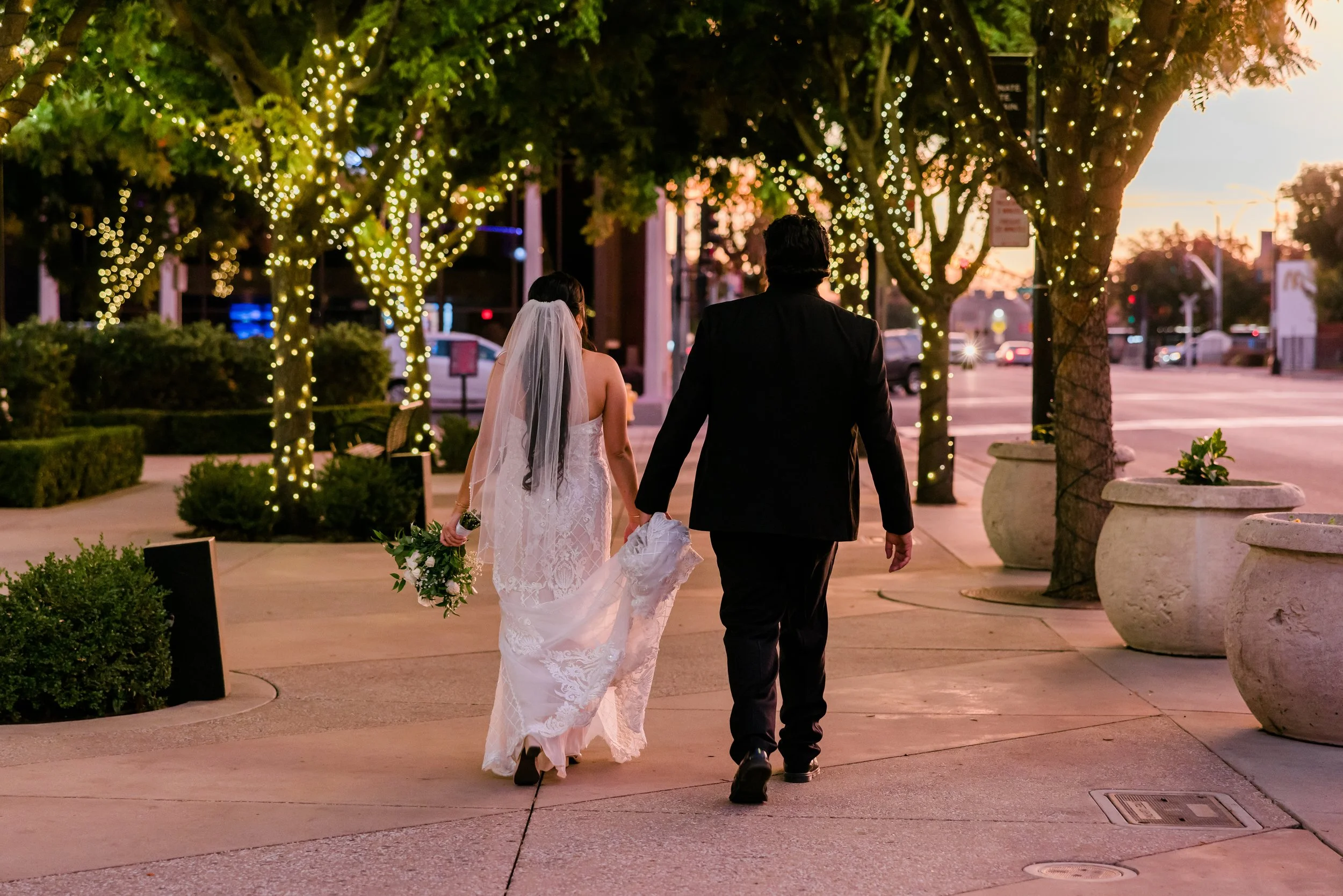 A newlywed couple walking hand-in-hand on a decorated street during sunset, with trees wrapped in string lights and large potted plants along the sidewalk.