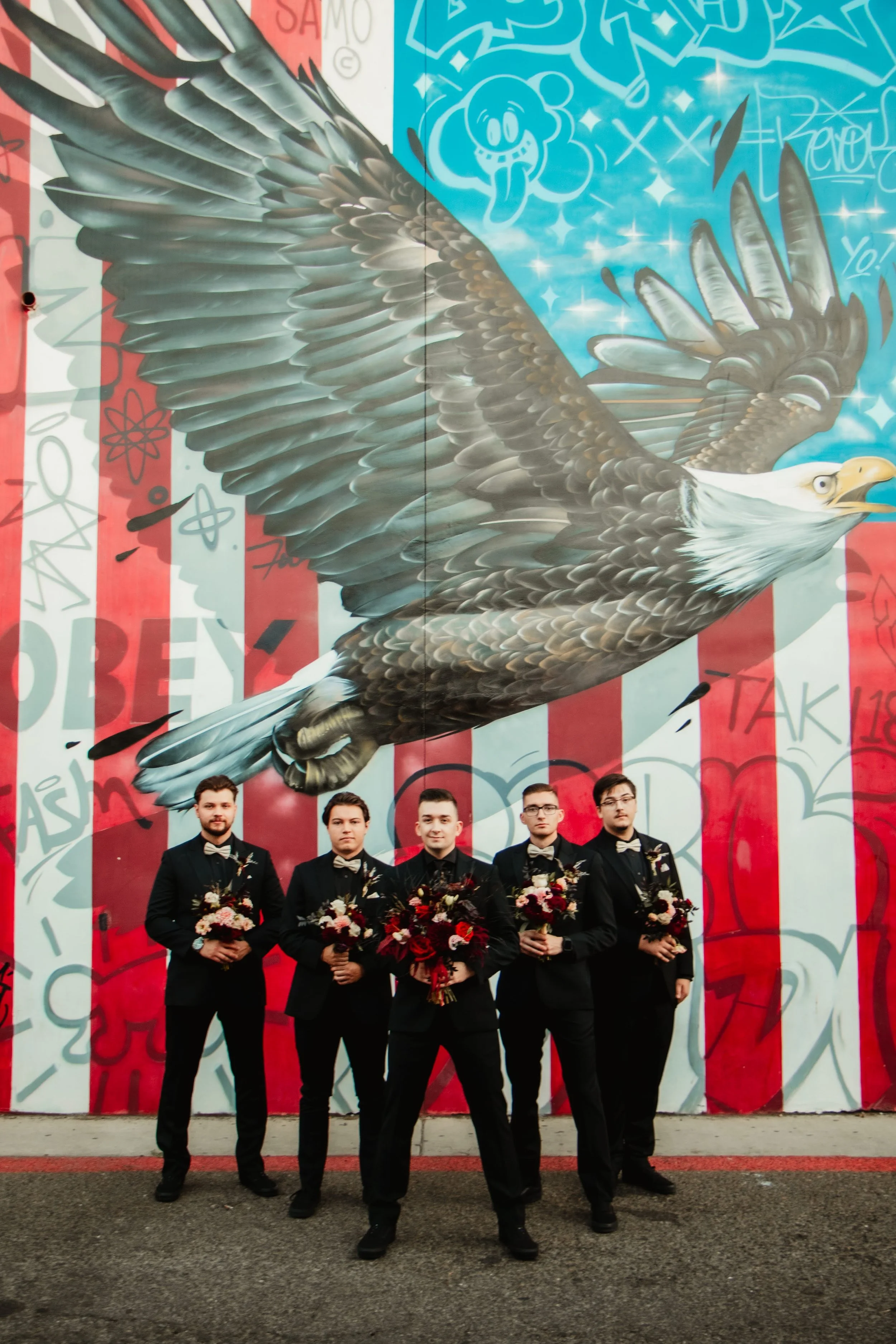 Five men in black suits and bow ties standing in front of a large mural of an eagle with outstretched wings, painted on a graffitied wall.