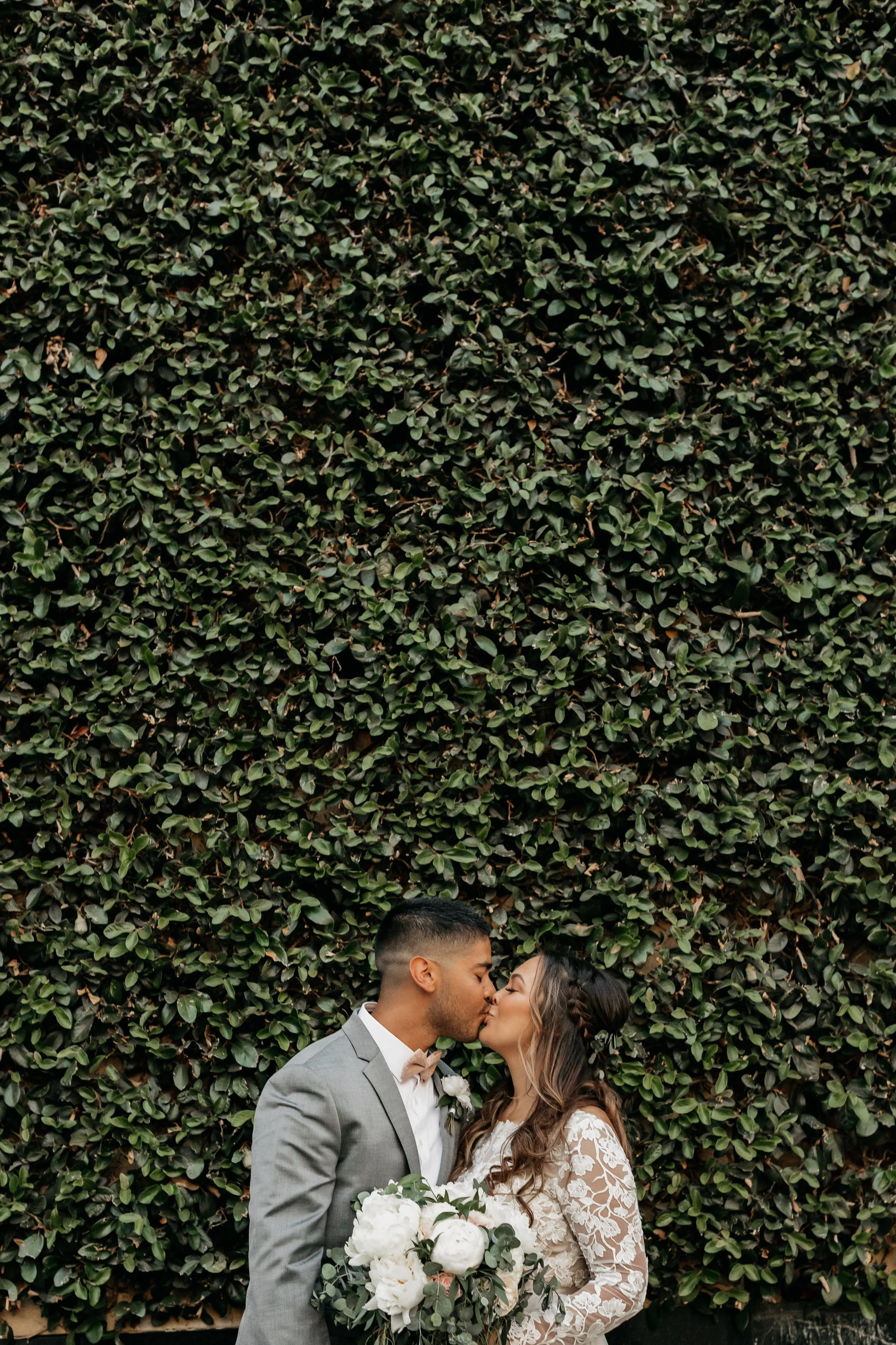 A newlywed couple sharing a kiss in front of a dense green hedge. The groom is dressed in a gray suit and beige bow tie, holding a bouquet of white peonies and greenery. The bride has long, wavy hair with braids and a lace wedding dress with floral p