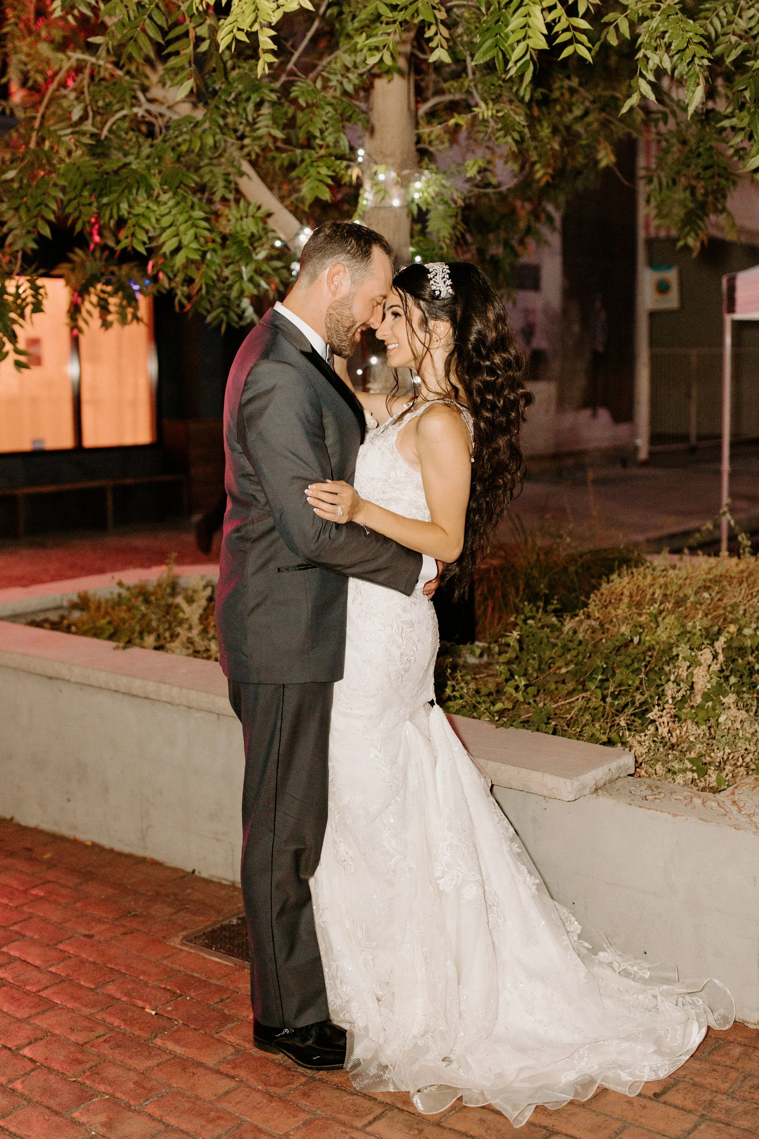 A bride and groom sharing an intimate moment outdoors at night before their wedding. The bride is wearing a white lace wedding dress with a train, and the groom is dressed in a dark suit. They are standing close, touching foreheads, and smiling. Ther