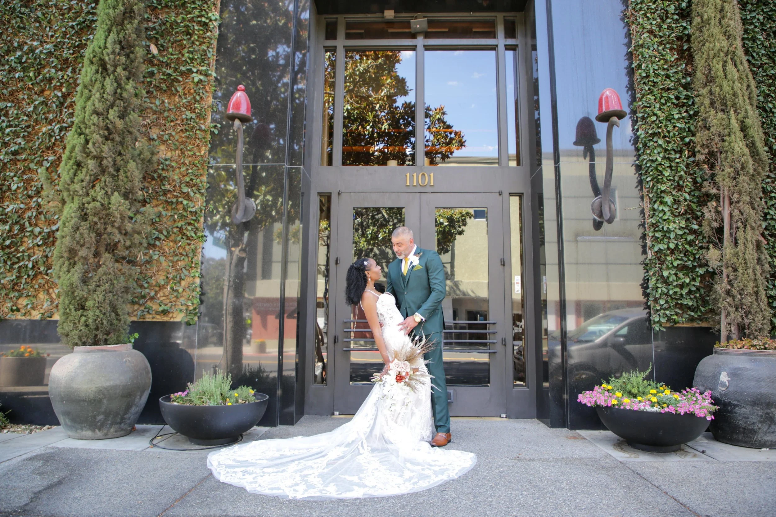A bride and groom standing close in front of a building entrance with the number 1101, surrounded by potted plants and wall decorations.