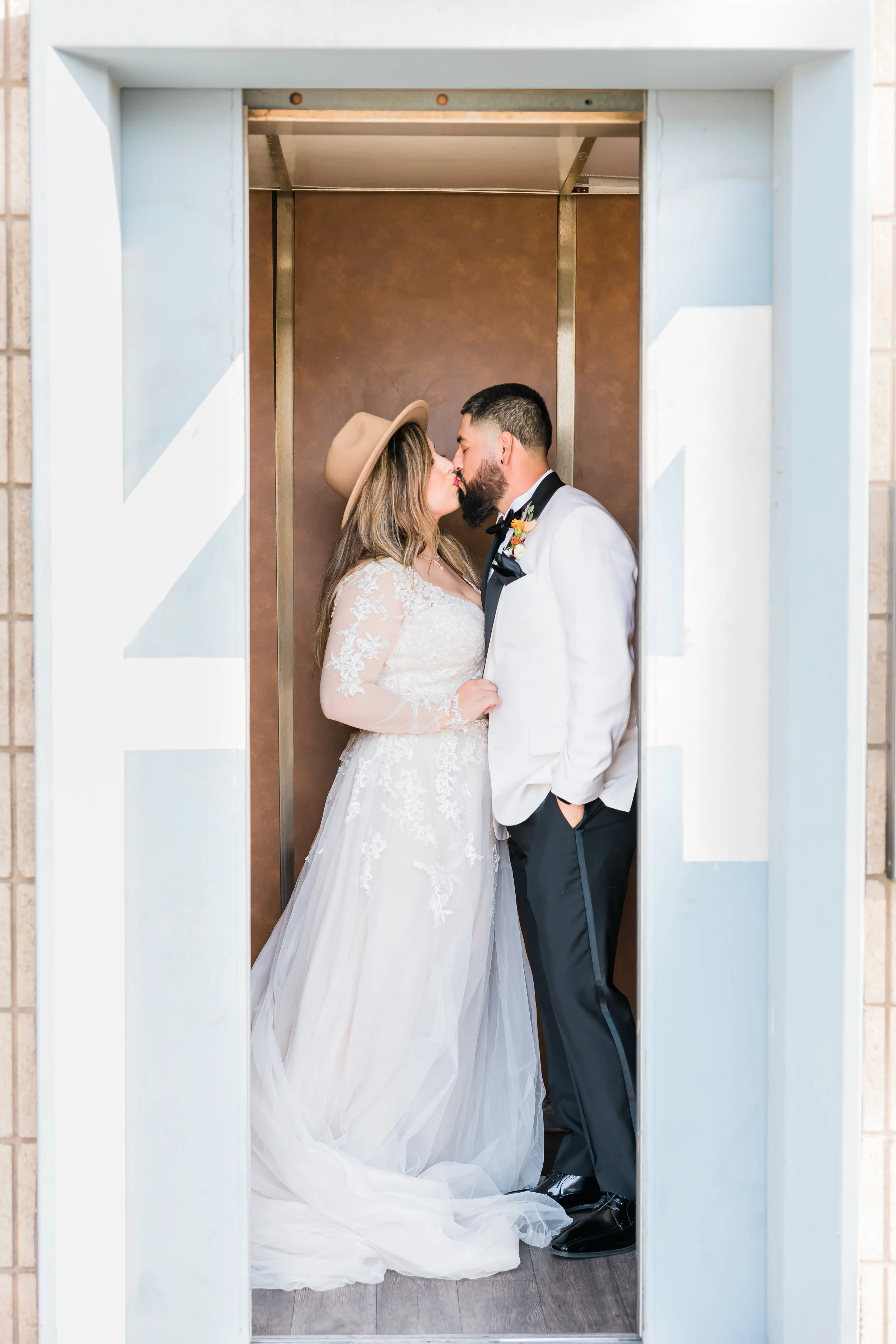 Bride and groom sharing a kiss in an elevator, the bride wearing a lace wedding dress and tan hat, and the groom in a white tuxedo jacket with black pants and bow tie.