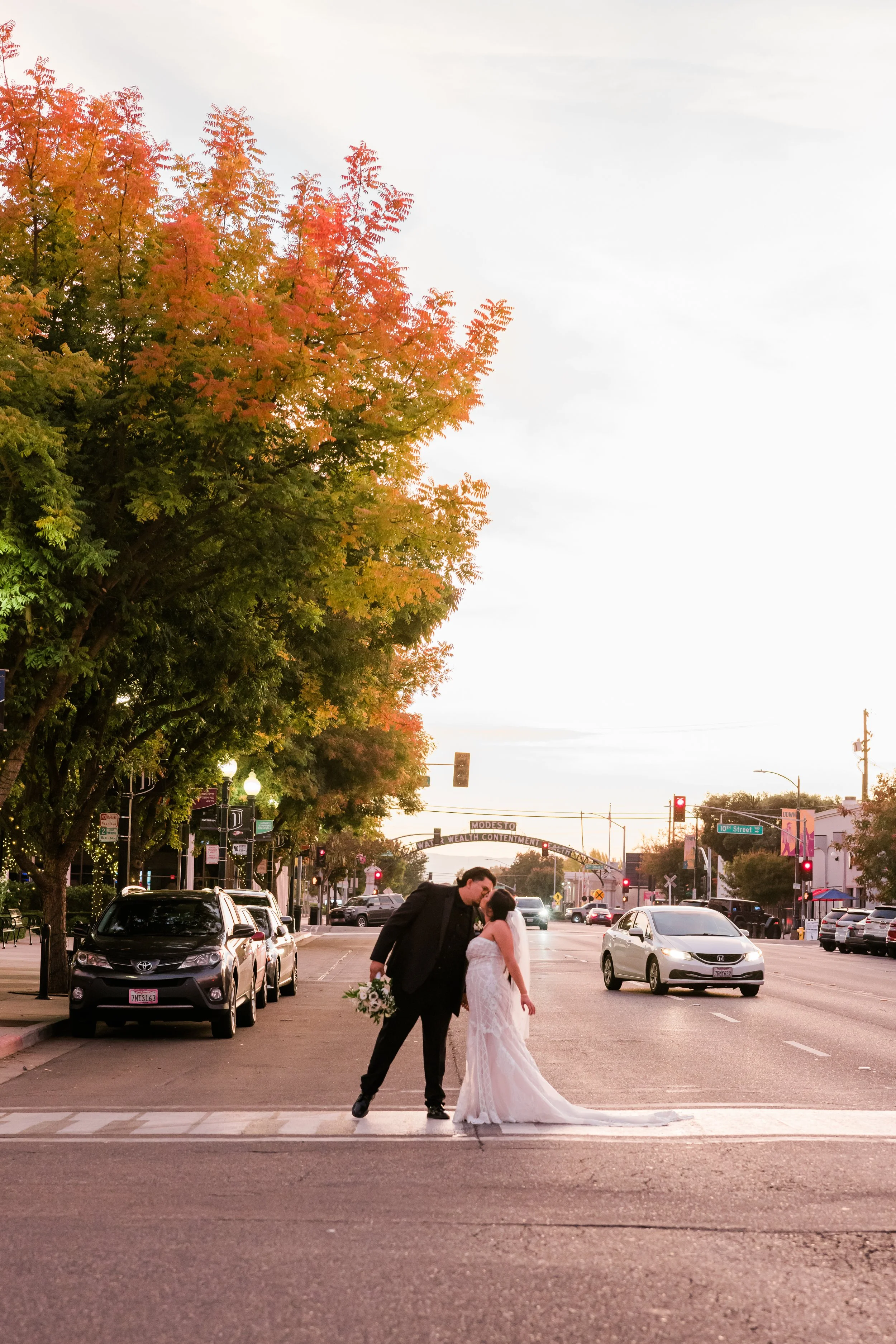 A newlywed couple sharing a kiss on a city street at sunset, with autumn trees and parked cars in the background.