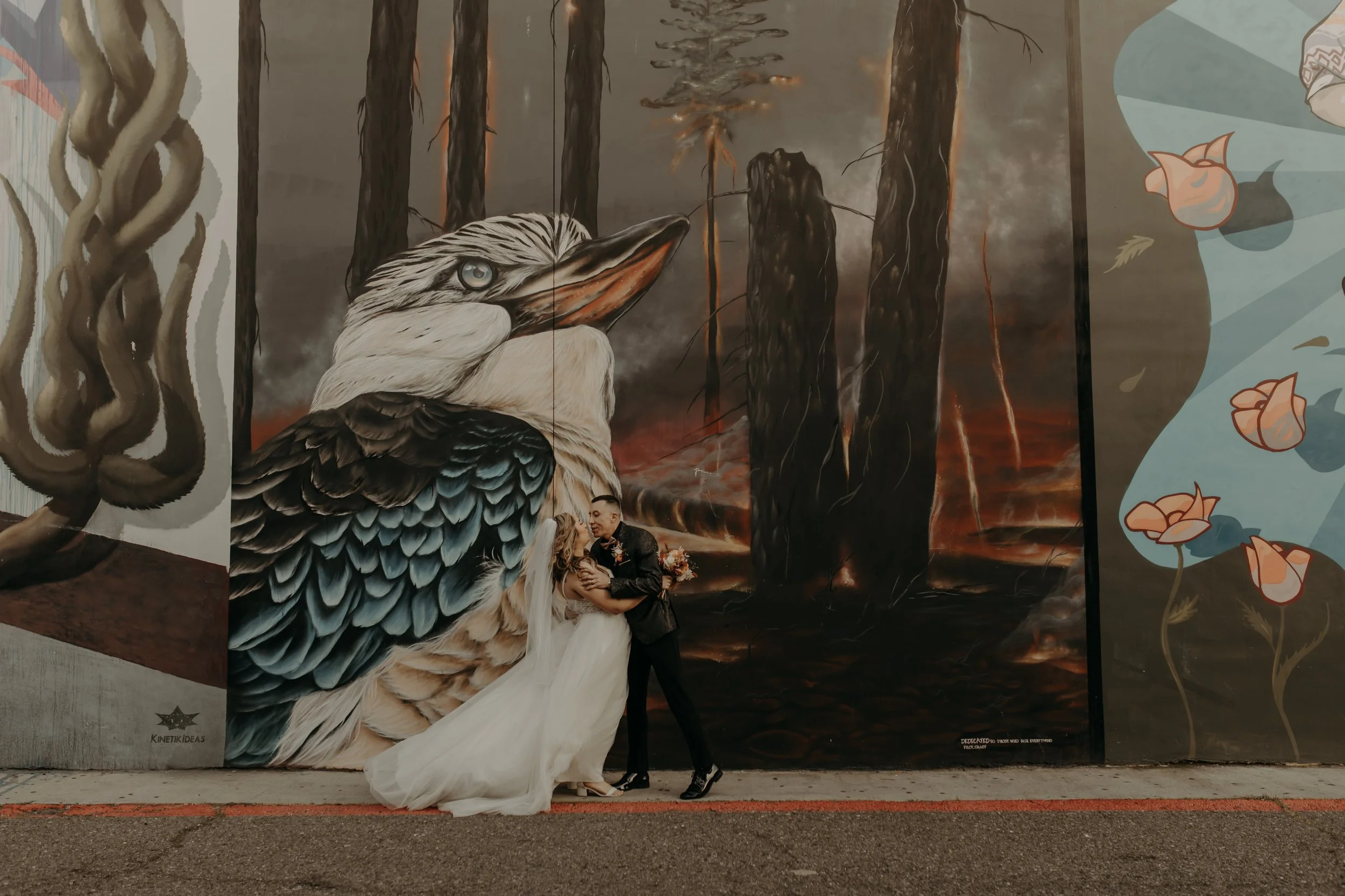 A couple in wedding attire embracing in front of a large mural featuring a detailed bird, possibly a hawk or falcon, against a stormy, fiery background and abstract floral designs on either side.