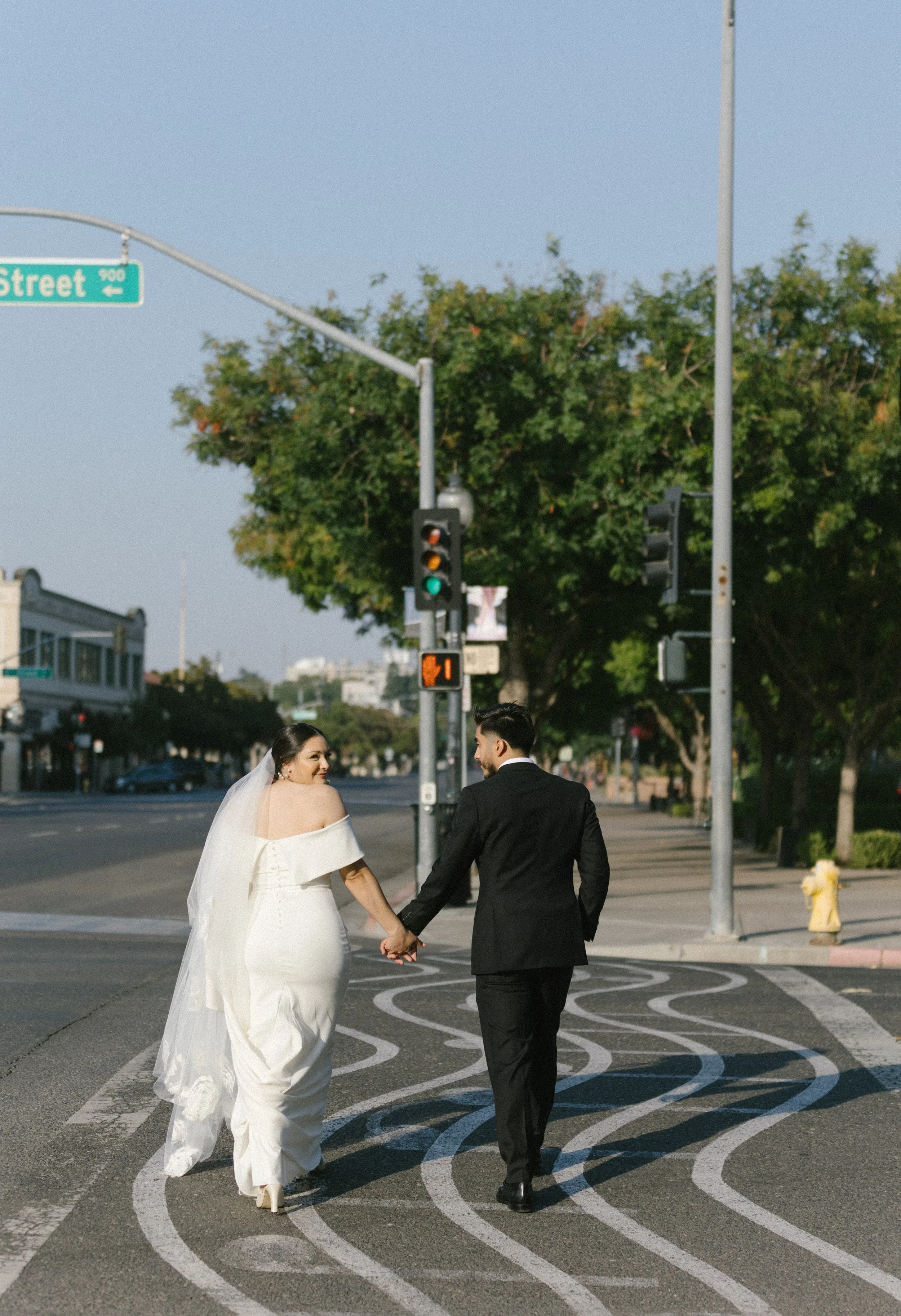 Bride and groom holding hands and walking across an empty city street at sunset, with trees lining the sidewalk and traffic lights overhead.