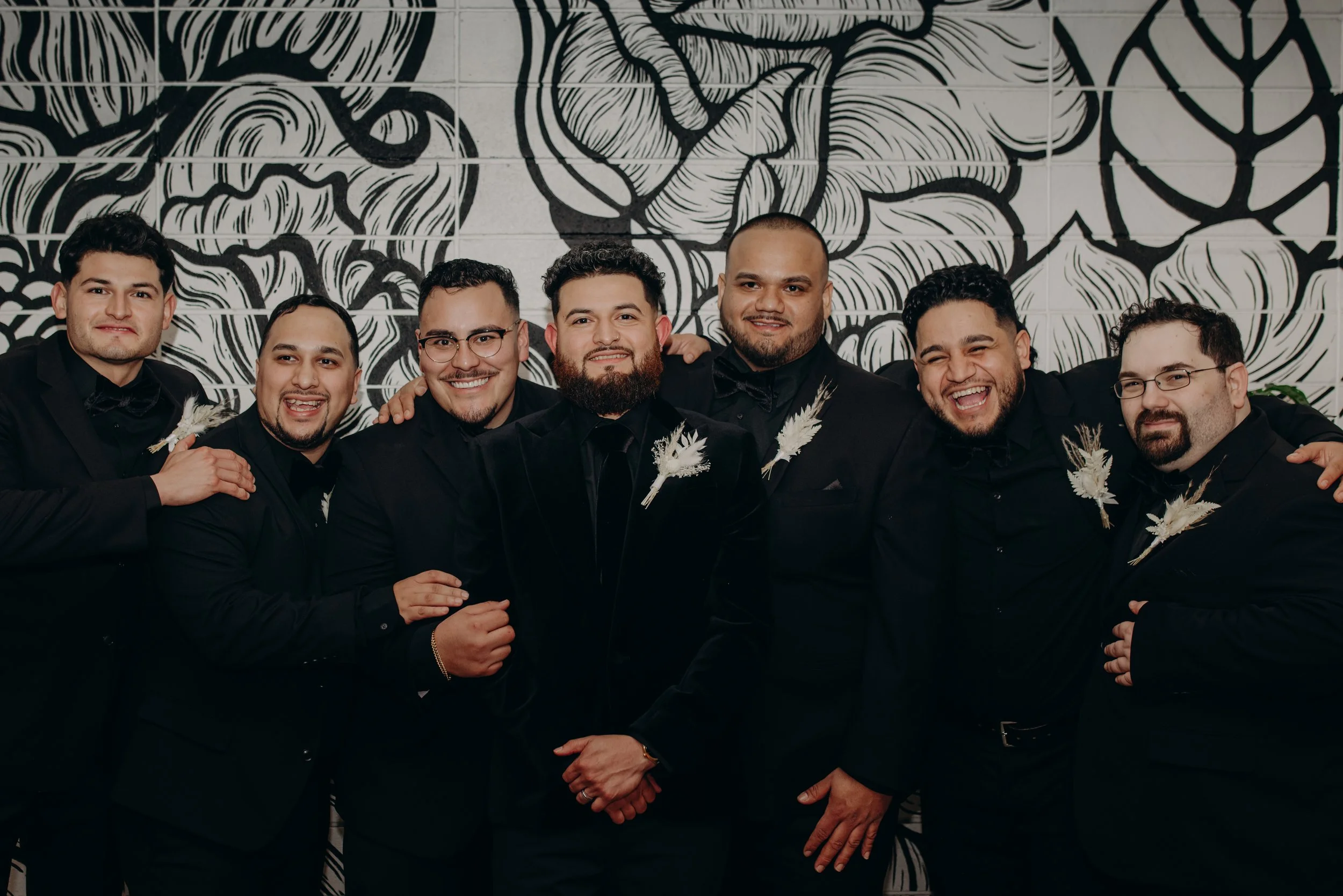 Group of seven men dressed in black suits, smiling and posing together in front of a black and white floral mural. Some have boutonnieres with white flowers and pampas grass.