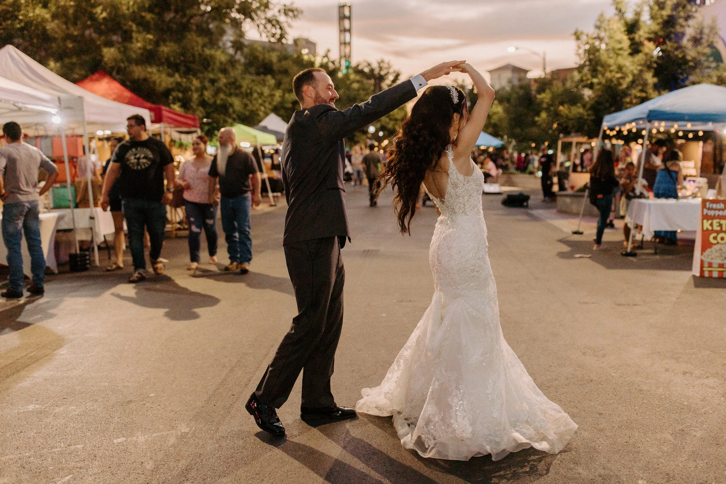 A couple dancing at an outdoor market during sunset. The woman is wearing a white wedding gown with lace details. The man is in a black suit. There are vendor tents in the background with people browsing.