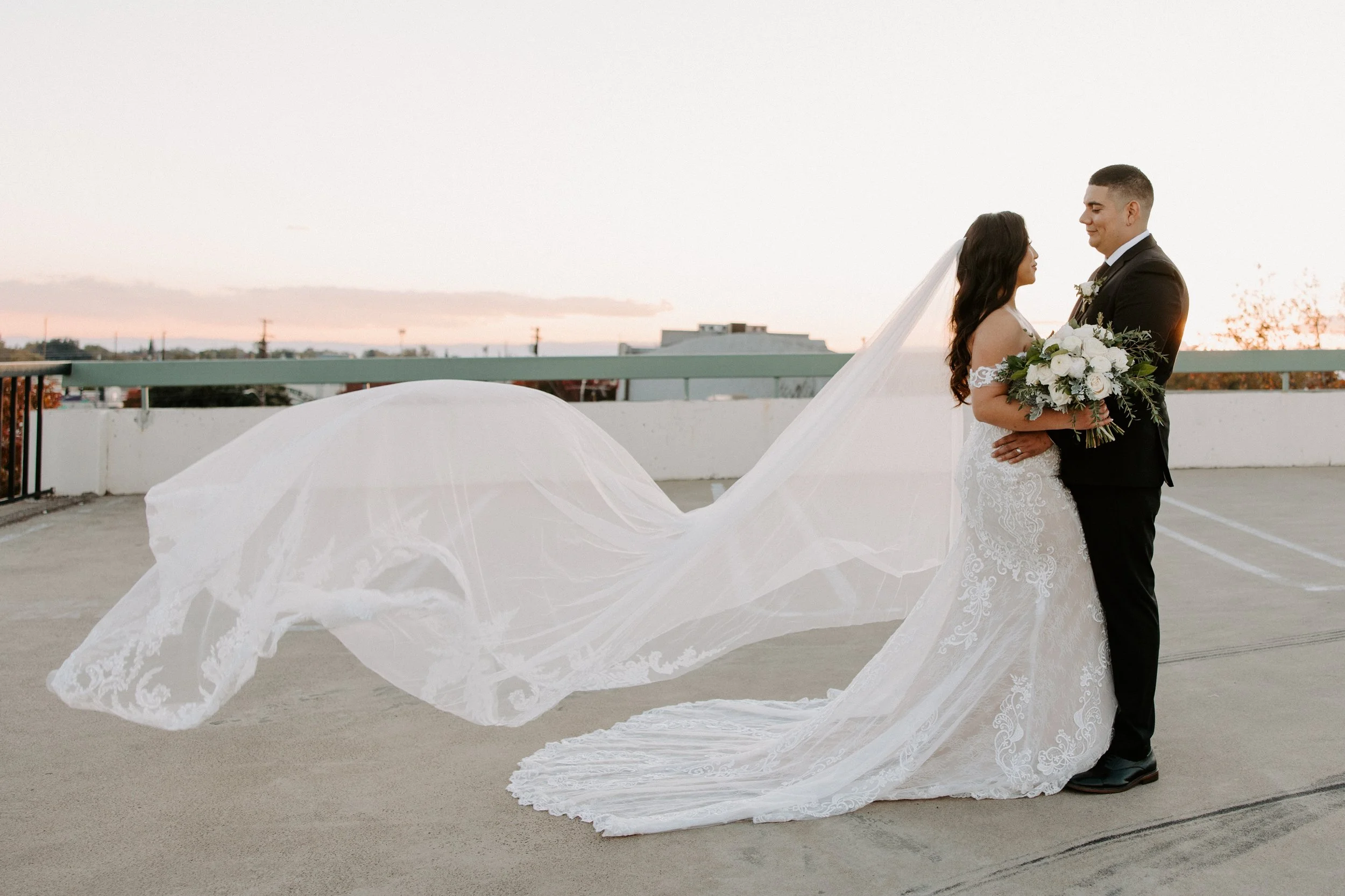 A bride and groom stand on a rooftop at sunset, holding a bouquet, with the bride's long veil flowing behind her.