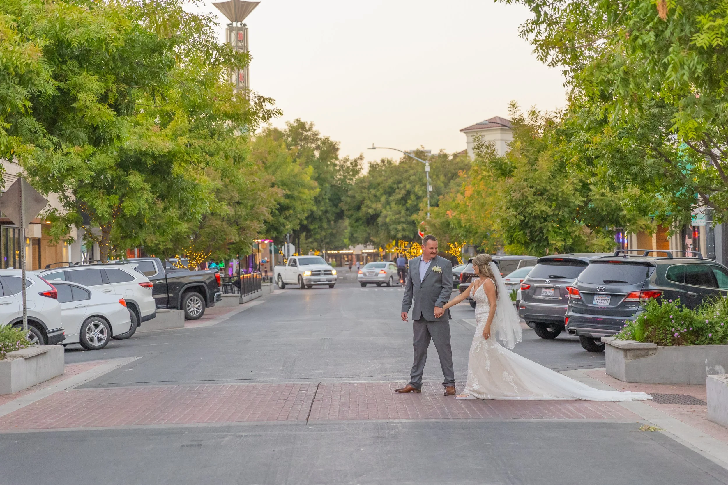 Bride and groom holding hands on a city street lined with parked cars and leafy trees, celebrating their wedding day.