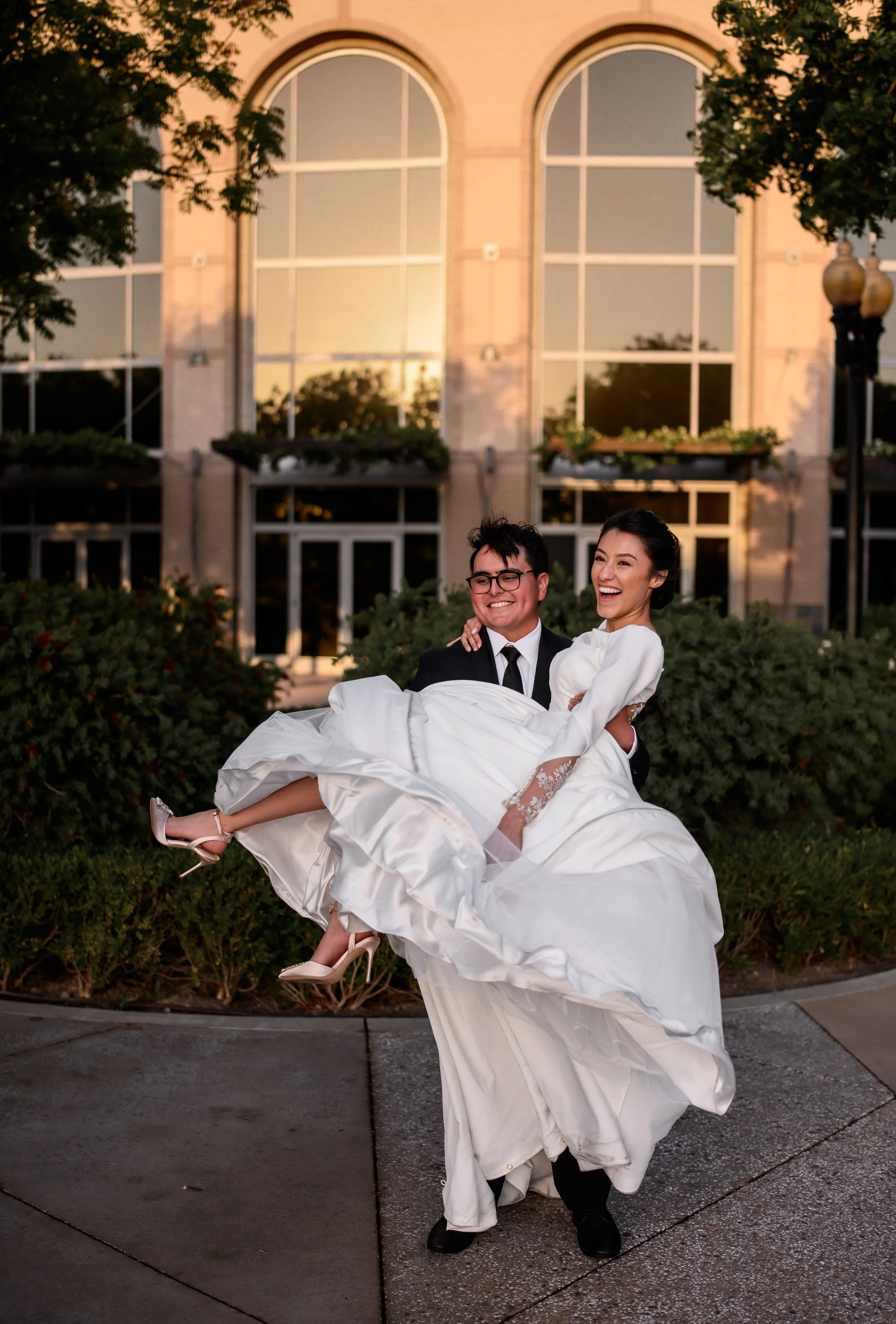 A smiling bride in a white wedding dress and heels being carried by a groom in a black suit, outside in front of a building at sunset.