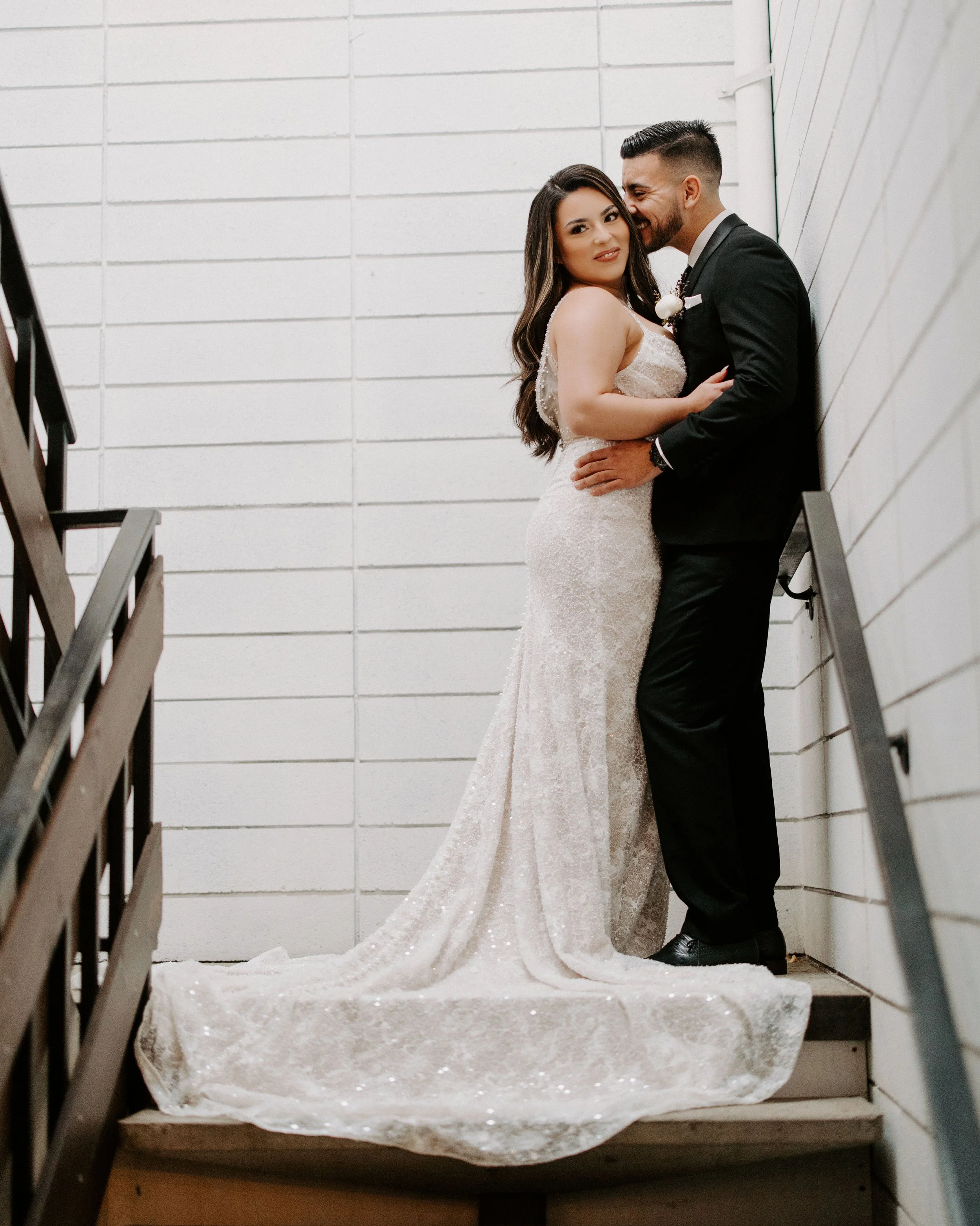 A bride and groom standing on a staircase, embracing and smiling, with white brick walls in the background.