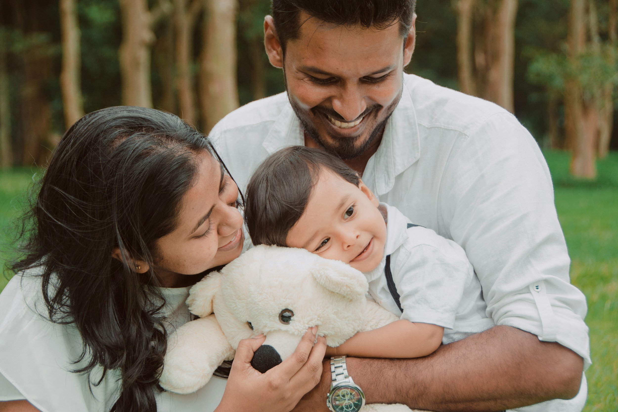 A happy family of three outdoors, hugging and smiling, holding a teddy bear in front of trees and grass.