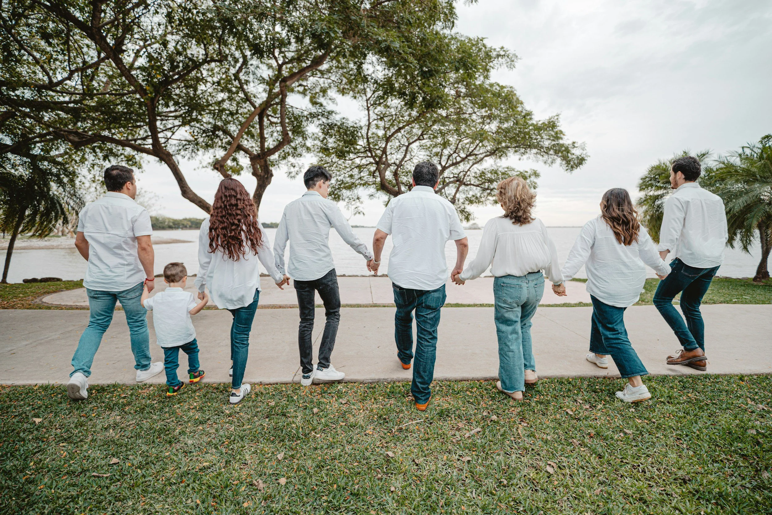 A group of eight people, including children, holding hands and walking along a lakeside park on a cloudy day.