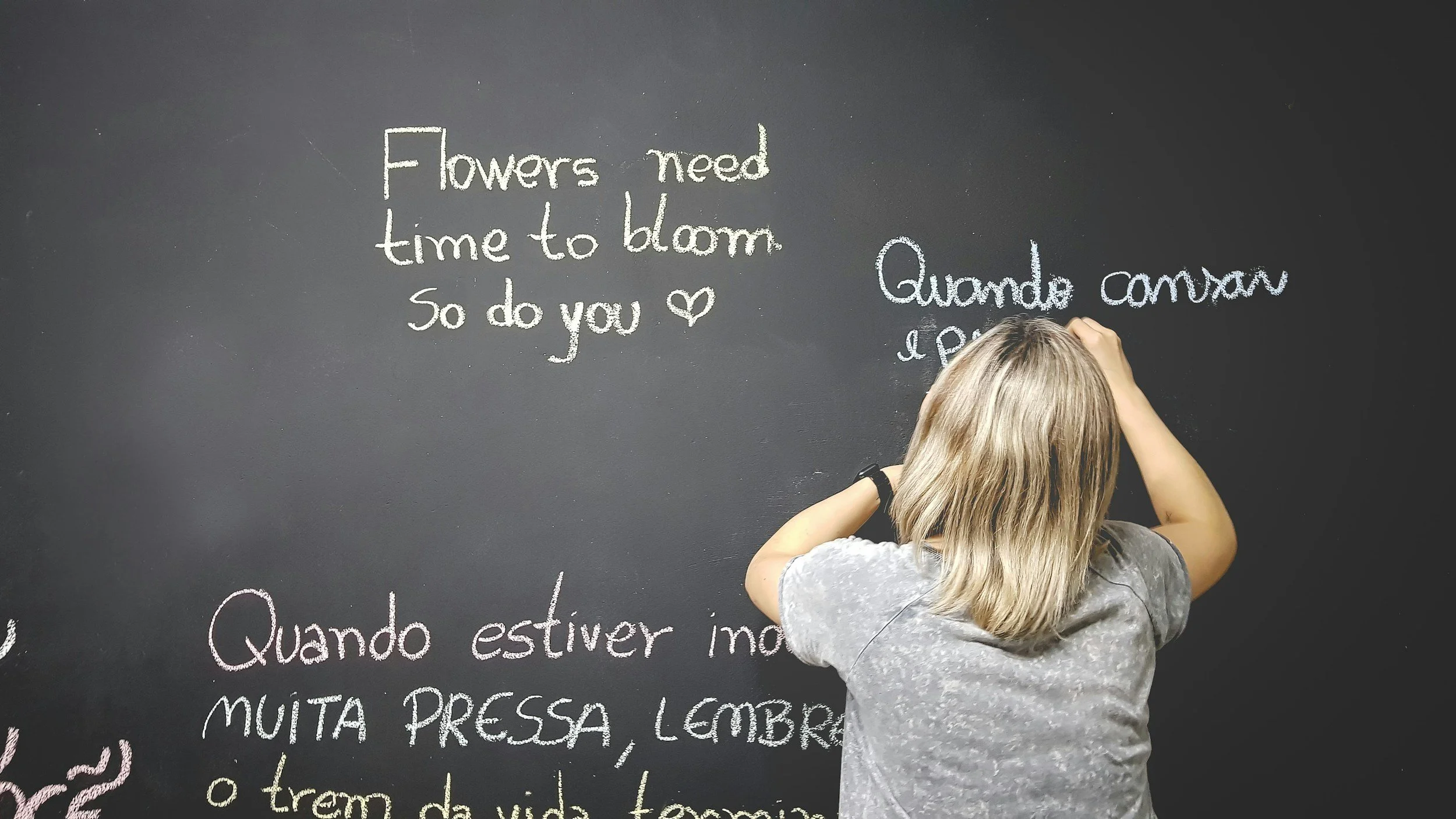 A person with blonde hair writing on a black chalkboard. The chalkboard has text written in different colors in Portuguese and English, about flowers and feelings.
