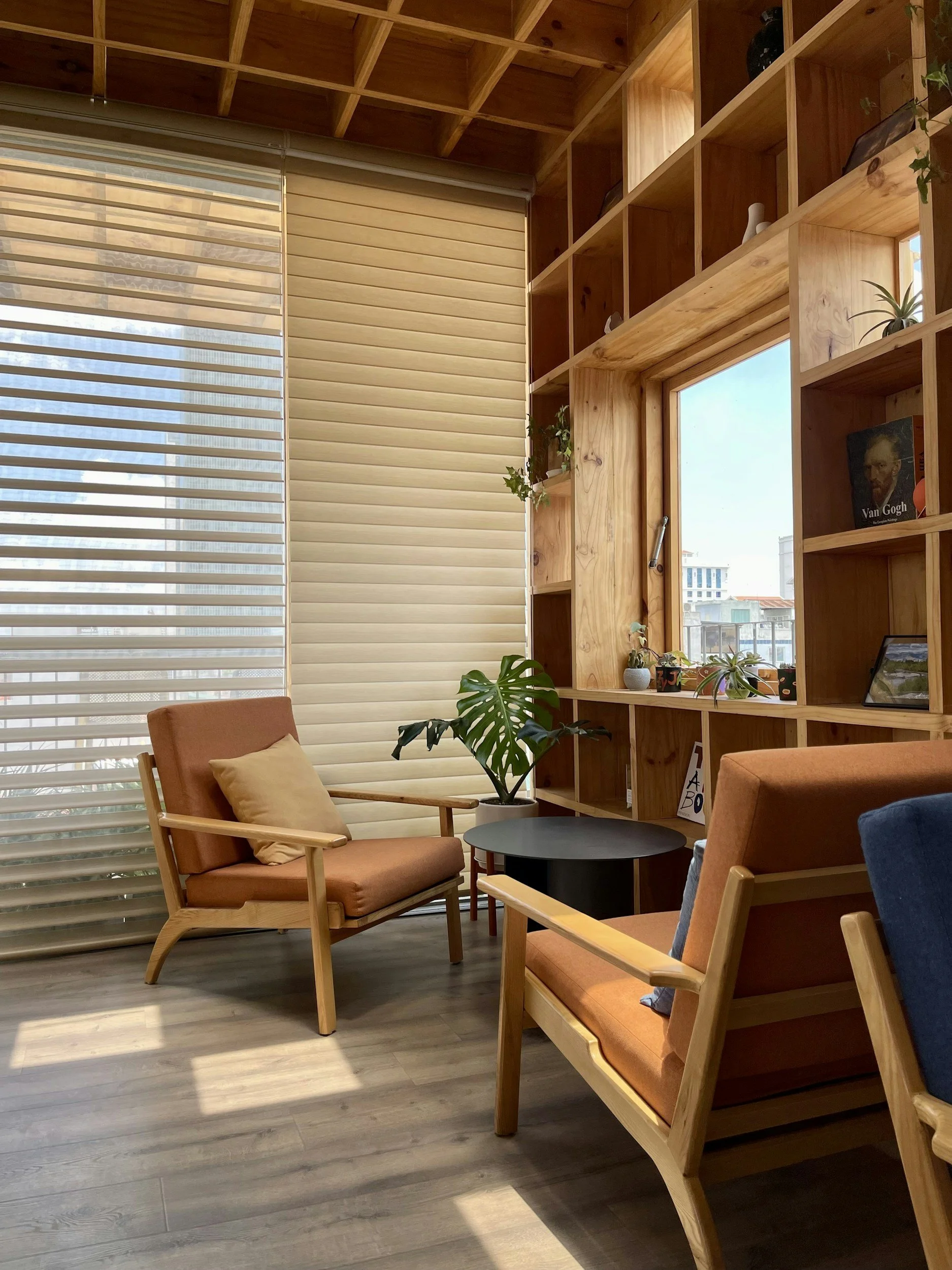 A cozy modern room with wooden furniture, a large window with blinds, and a wooden shelf built into the wall. The room includes two wooden armchairs with orange cushions, a small round black table, and indoor plants.