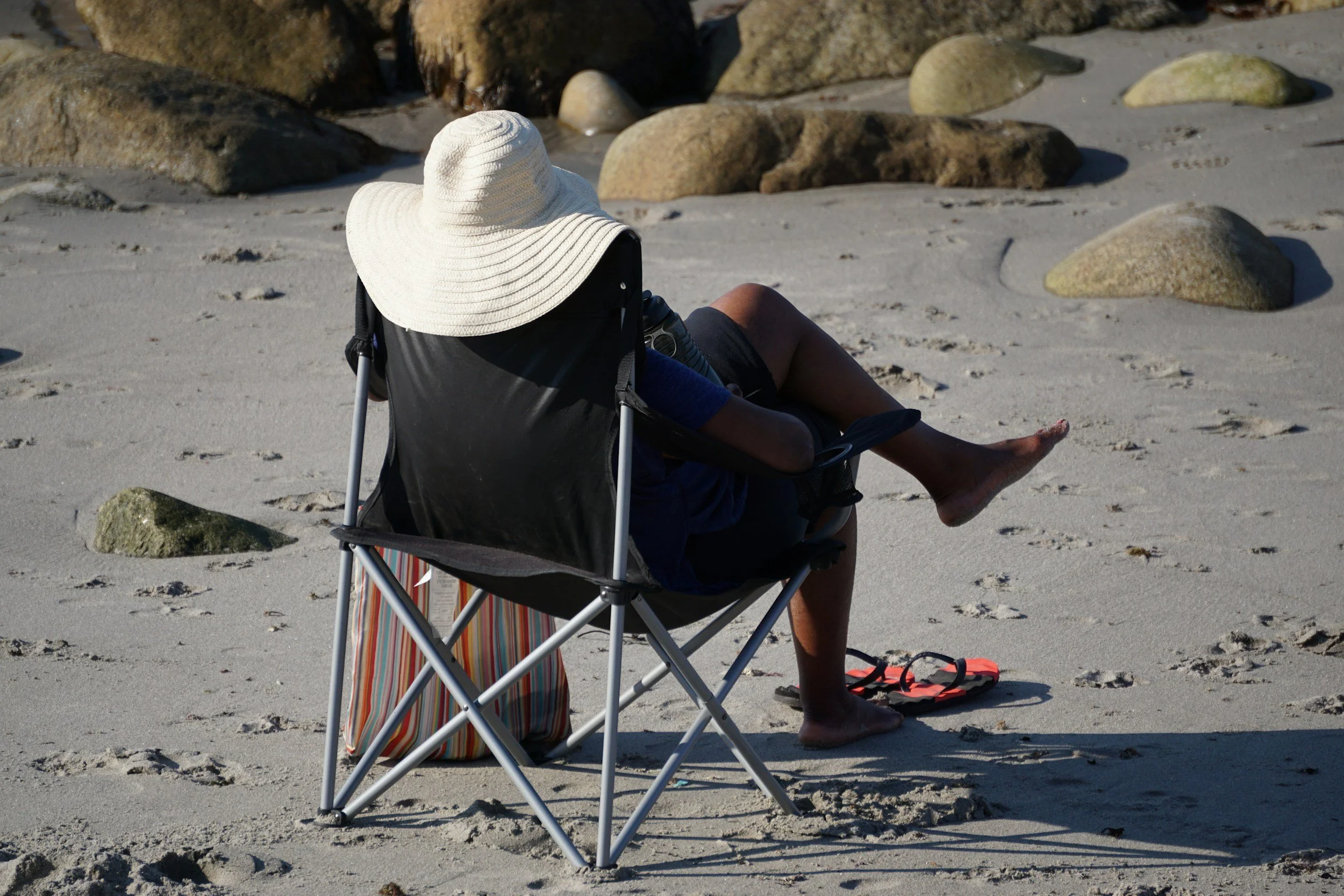 A person sitting in a portable chair on a sandy beach, wearing a large sun hat, with rocks in the background, and items like flip-flops and a striped bag nearby.