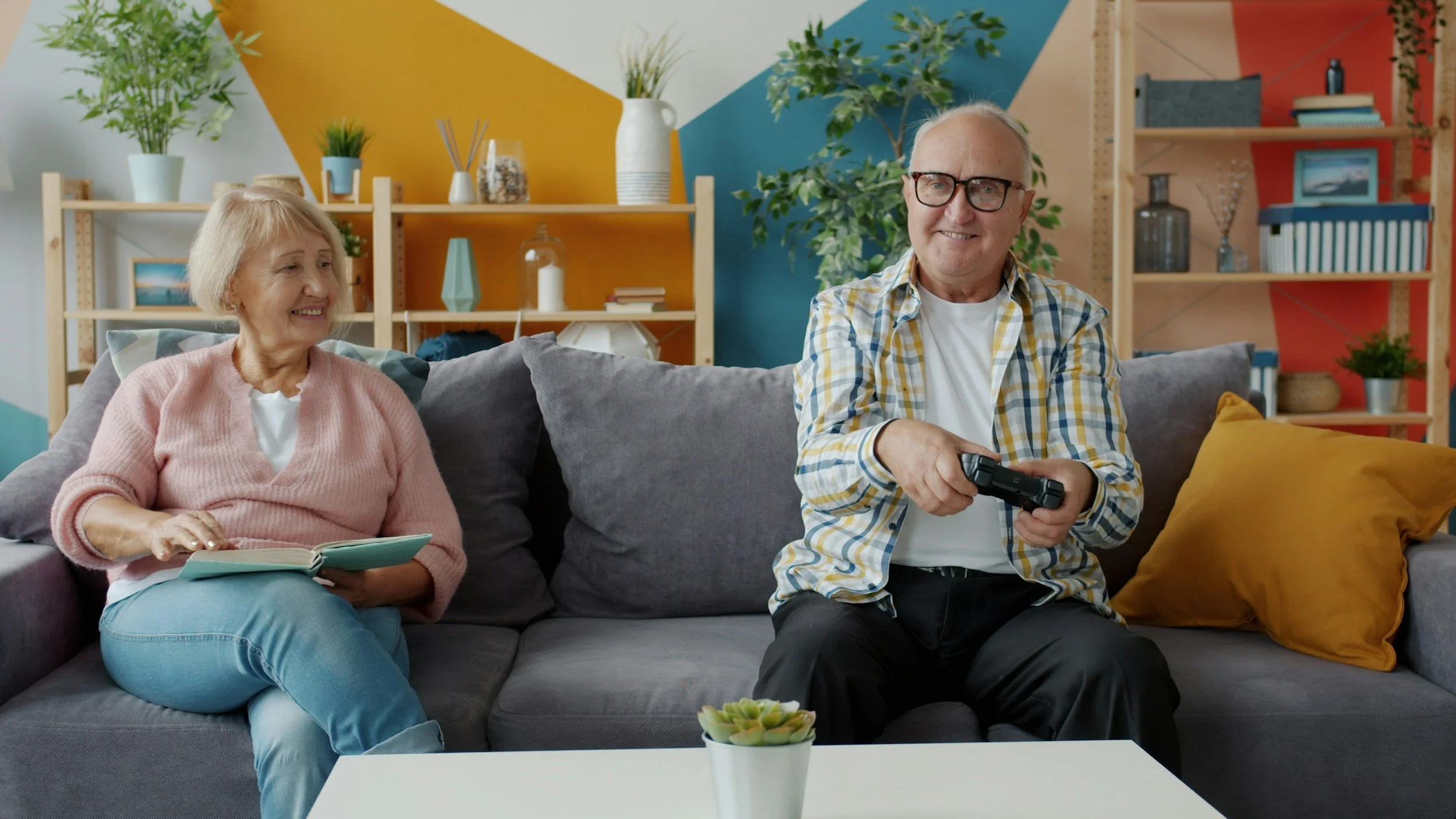 An elderly woman and man sitting on a gray sofa playing video games together. The woman holds a book, smiling, while the man holds a game controller, smiling. The living room has colorful geometric wall art, plants, shelves with decor, and a white table with a small plant in front.