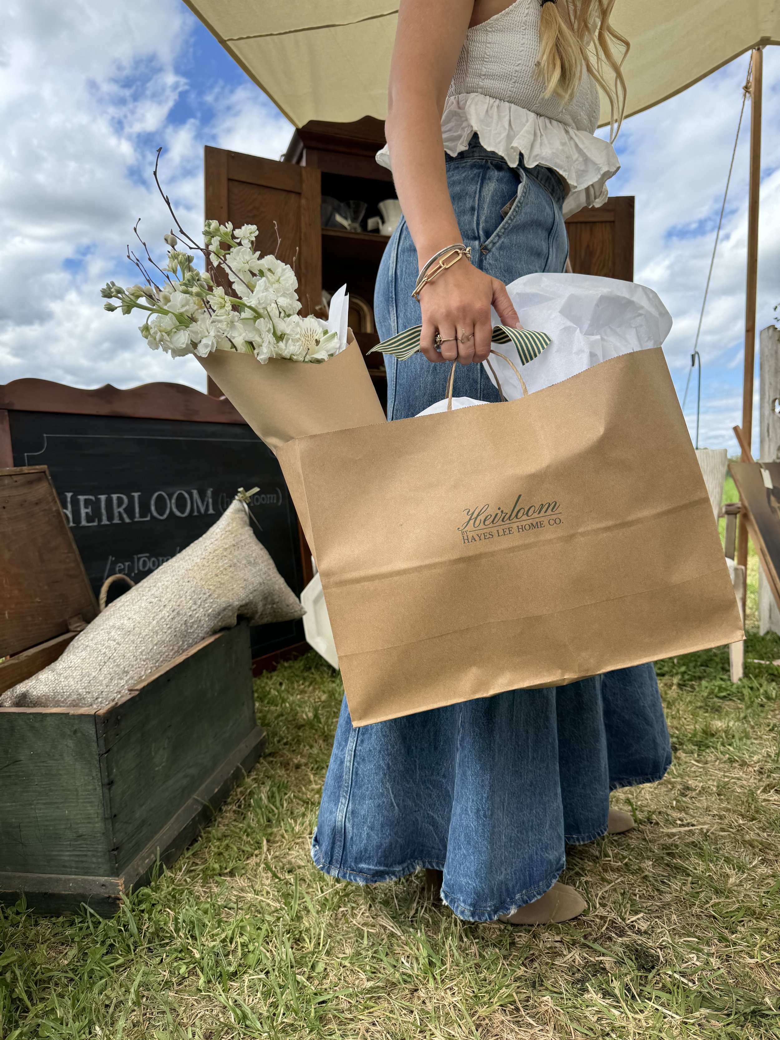 Person holding a brown shopping bag filled with wrapped items and white flowers, standing outdoors with a rustic setup in the background.