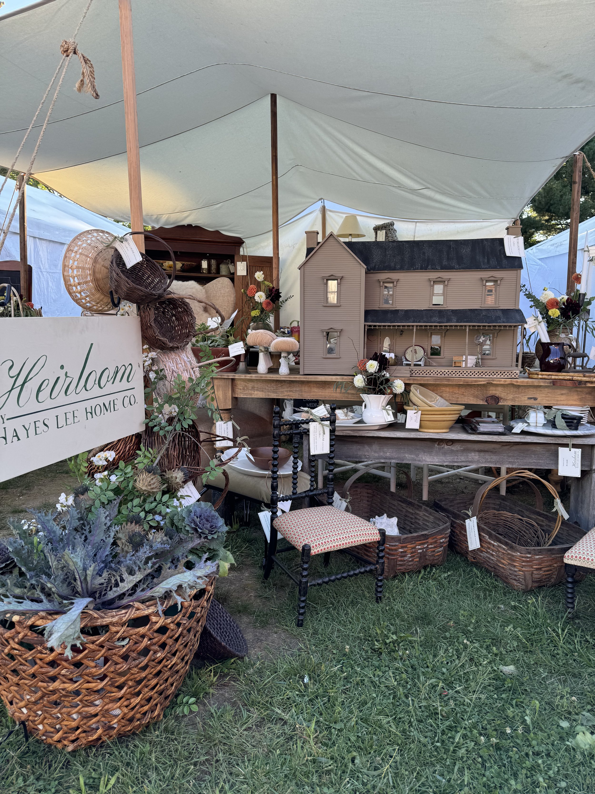 Outdoor market stall with vintage items, including a dollhouse, baskets, plants, woven hats, and a decorative chair. A sign reads 'Heirloom Hayes Lee Home Co.'