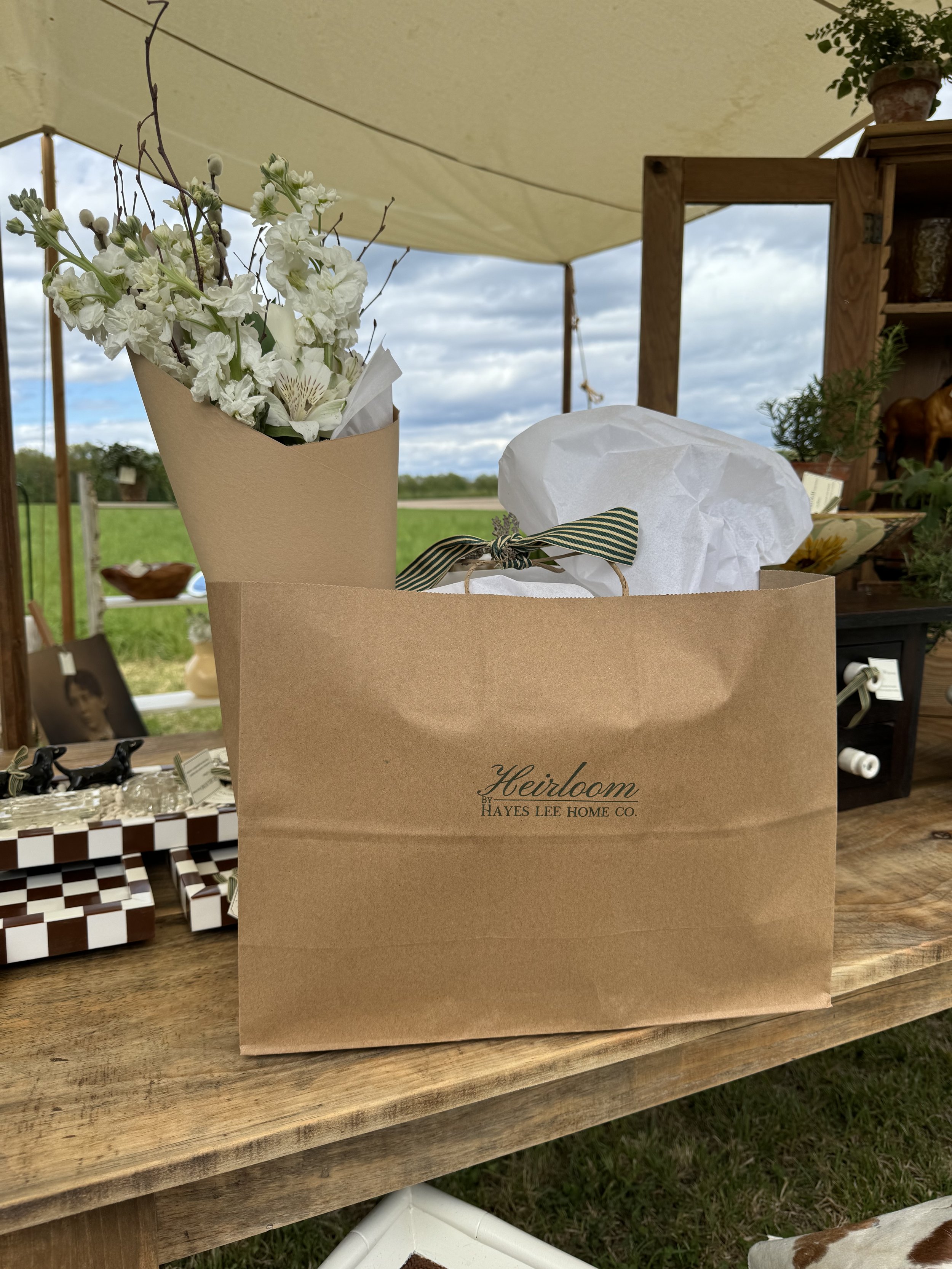 A wooden table with a brown paper bag labeled "Heirloom Hayes Lee Home Co." next to a bouquet of white flowers wrapped in brown paper and gift wrapped with tissue paper, under a tent canopy.