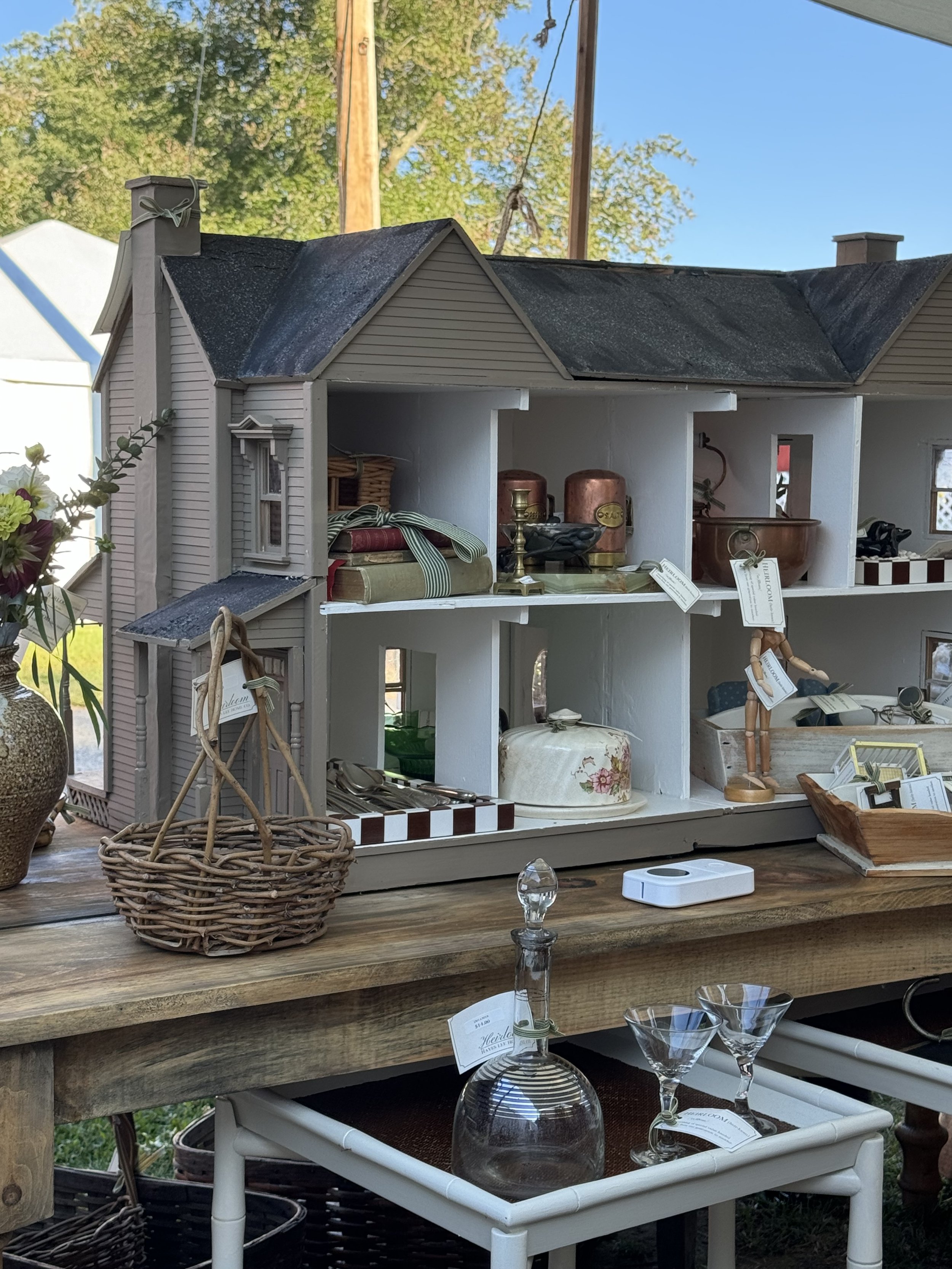 A dollhouse displayed on a wooden table, with various miniature objects inside. A wicker basket is on the table, alongside a glass decanter, martini glasses, and other decorative items. The setup suggests an antique or crafts fair setting.