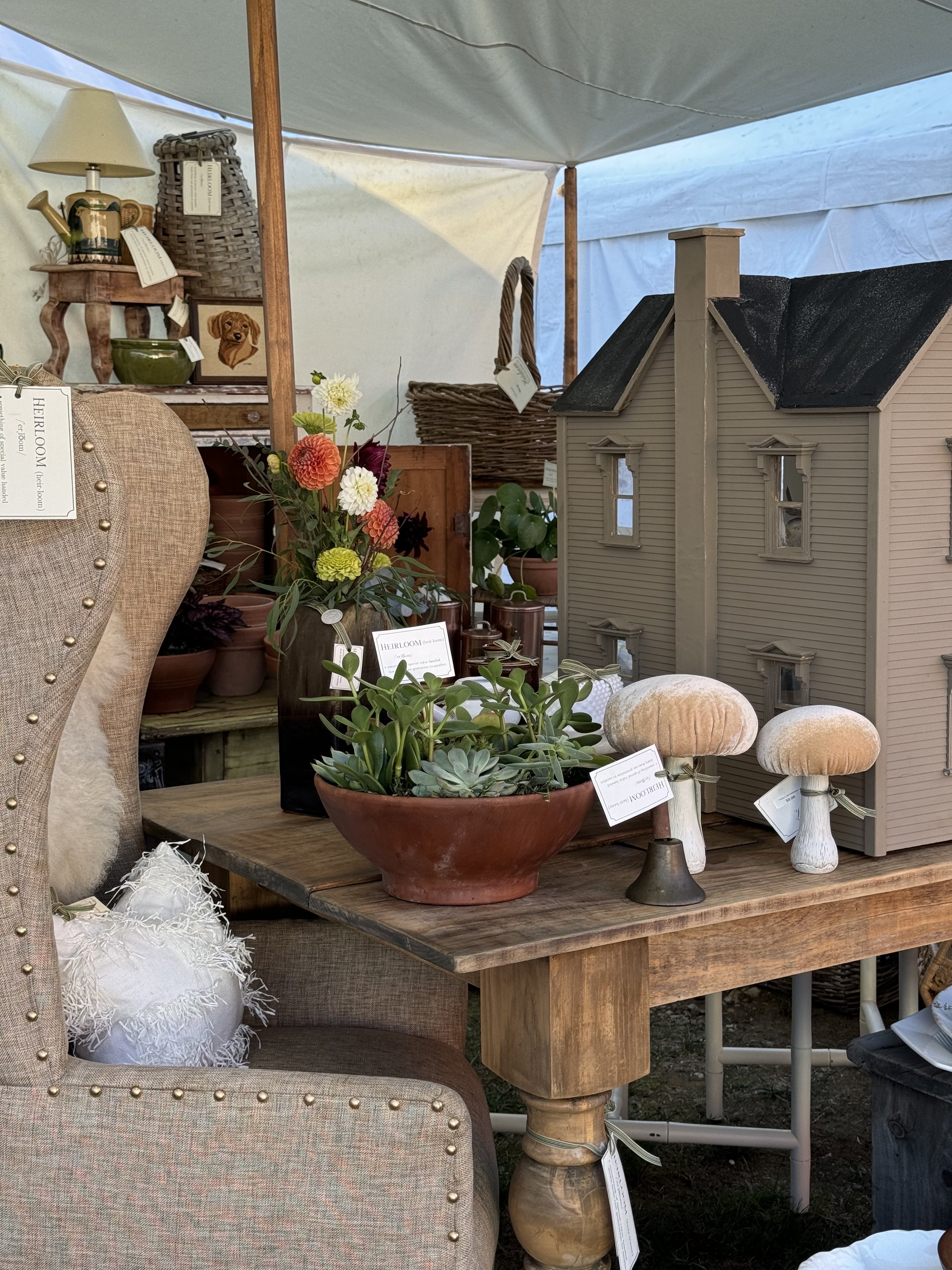 Indoor setup featuring a model dollhouse, a beige cushioned chair, a wooden table with potted plants and decorative mushrooms, a lamp, and decorative items. Displayed in a tent setting.
