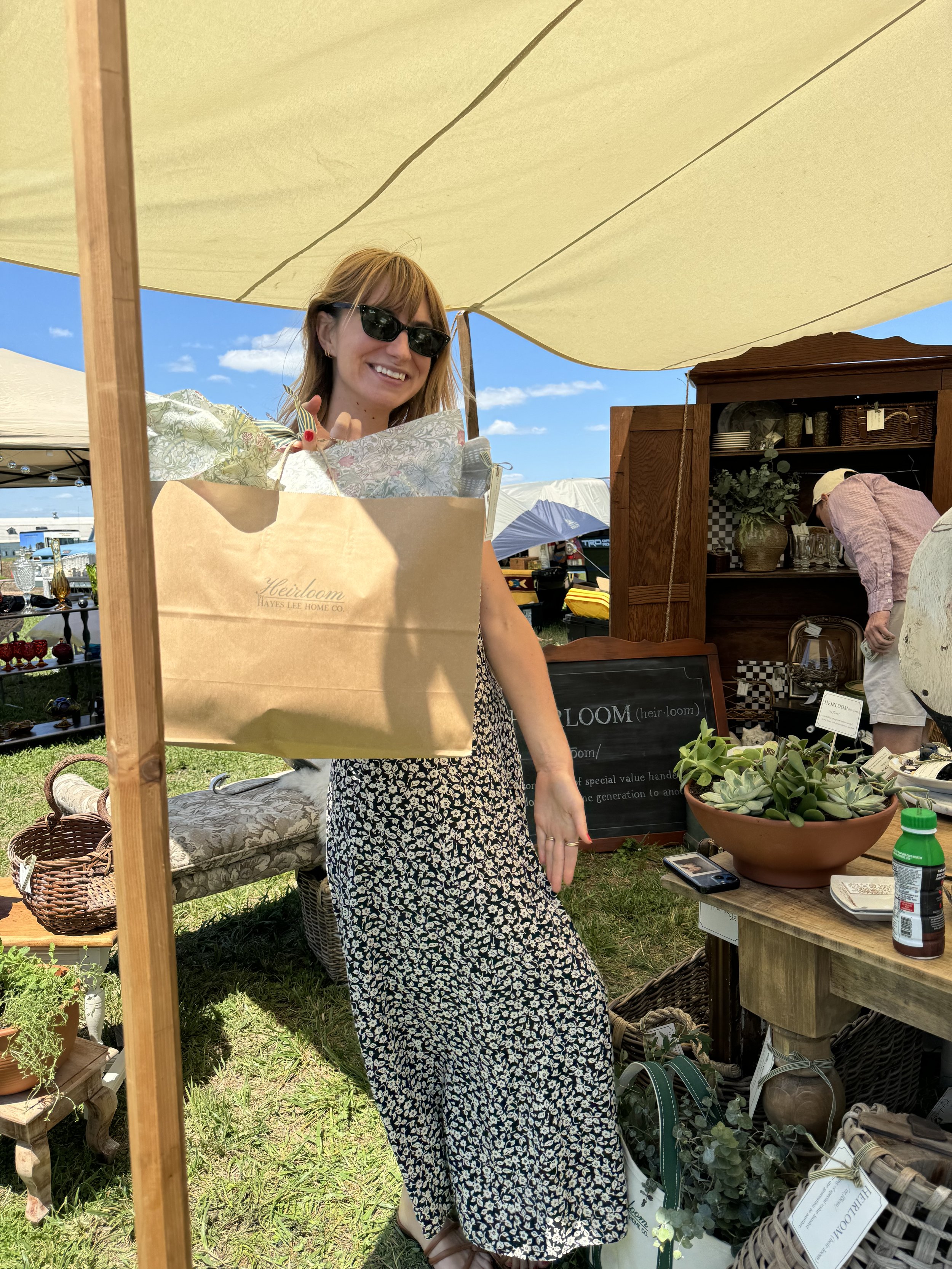 A woman smiling and holding a large shopping bag at an outdoor market. She is wearing a floral dress and sunglasses. The stall is decorated with plants, baskets, and home decor items. Another person is browsing in the background.