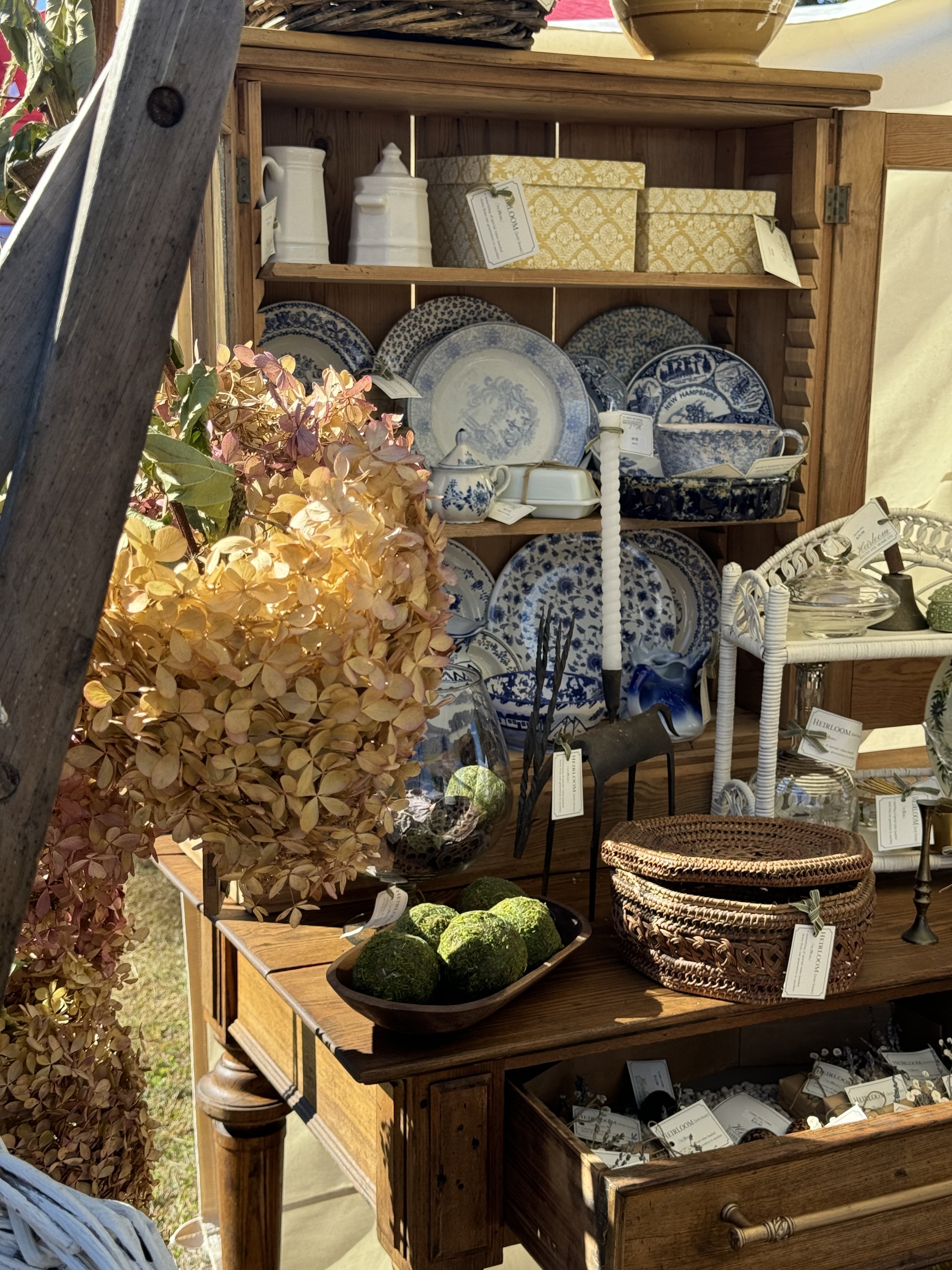 A wooden display cabinet with blue and white ceramic plates, bowls, and jugs. Dried hydrangeas, green moss balls, woven baskets, and decorative items are arranged around the cabinet.