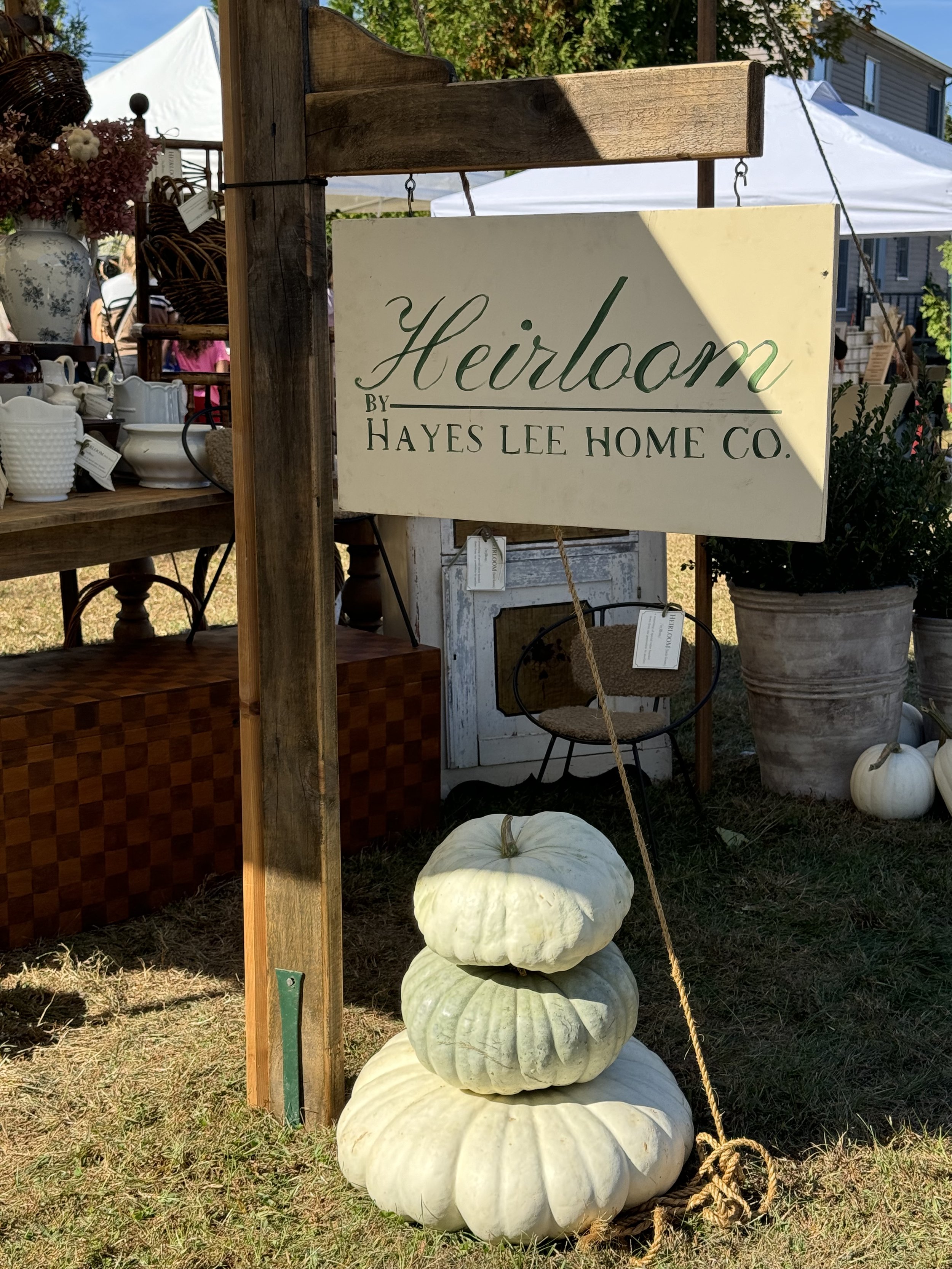 Outdoor market display with "Heirloom by Hayes Lee Home Co." sign, stacked white pumpkins, vintage items on a wooden table, and potted plants under a tent.