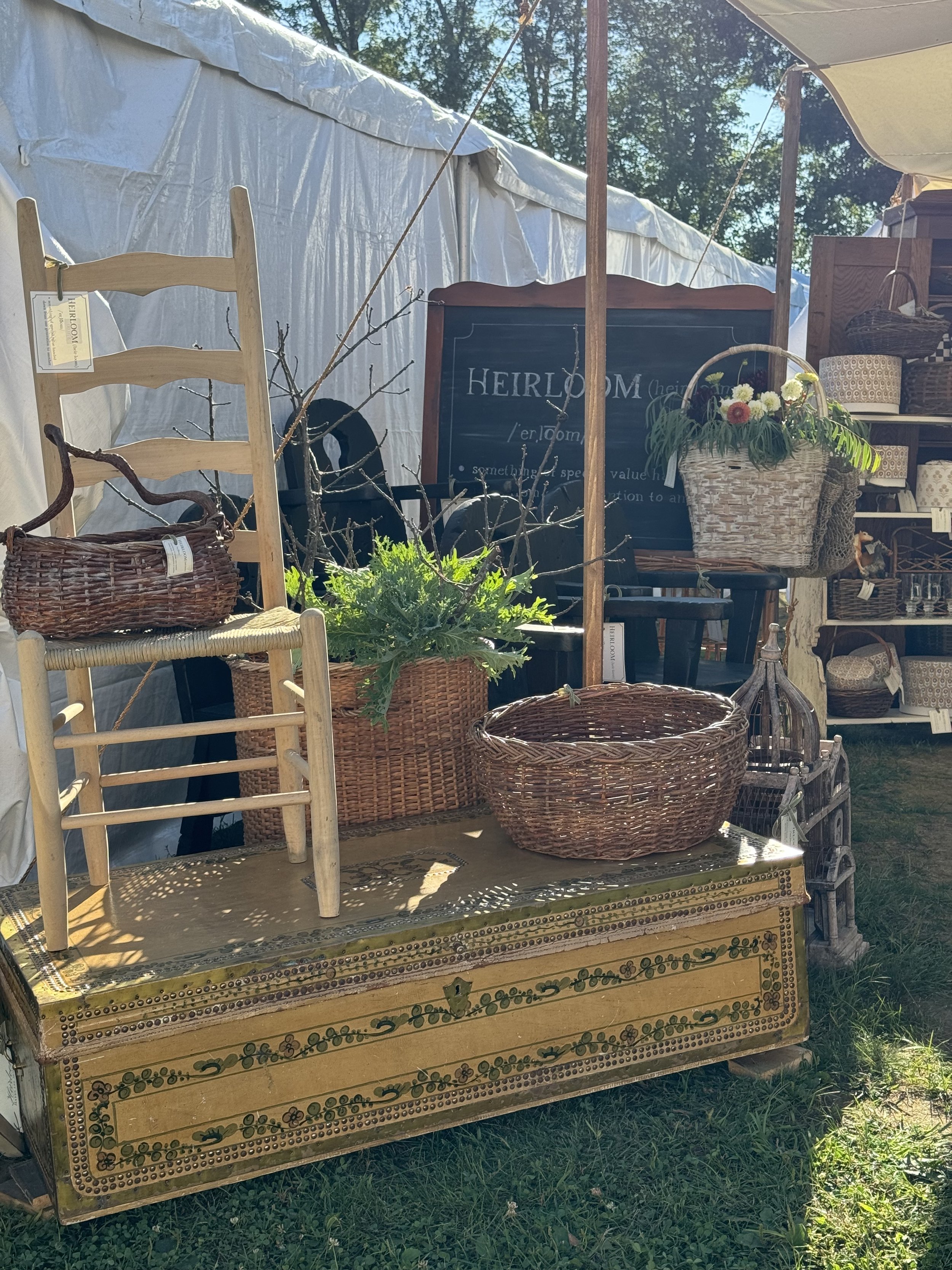 Outdoor display at a flea market with a wooden chair, wicker baskets, a painted chest, and greenery. A sign with the word "HEIRLOOM" is visible, along with a tent canopy overhead. Sunlight creates shadows on the items.